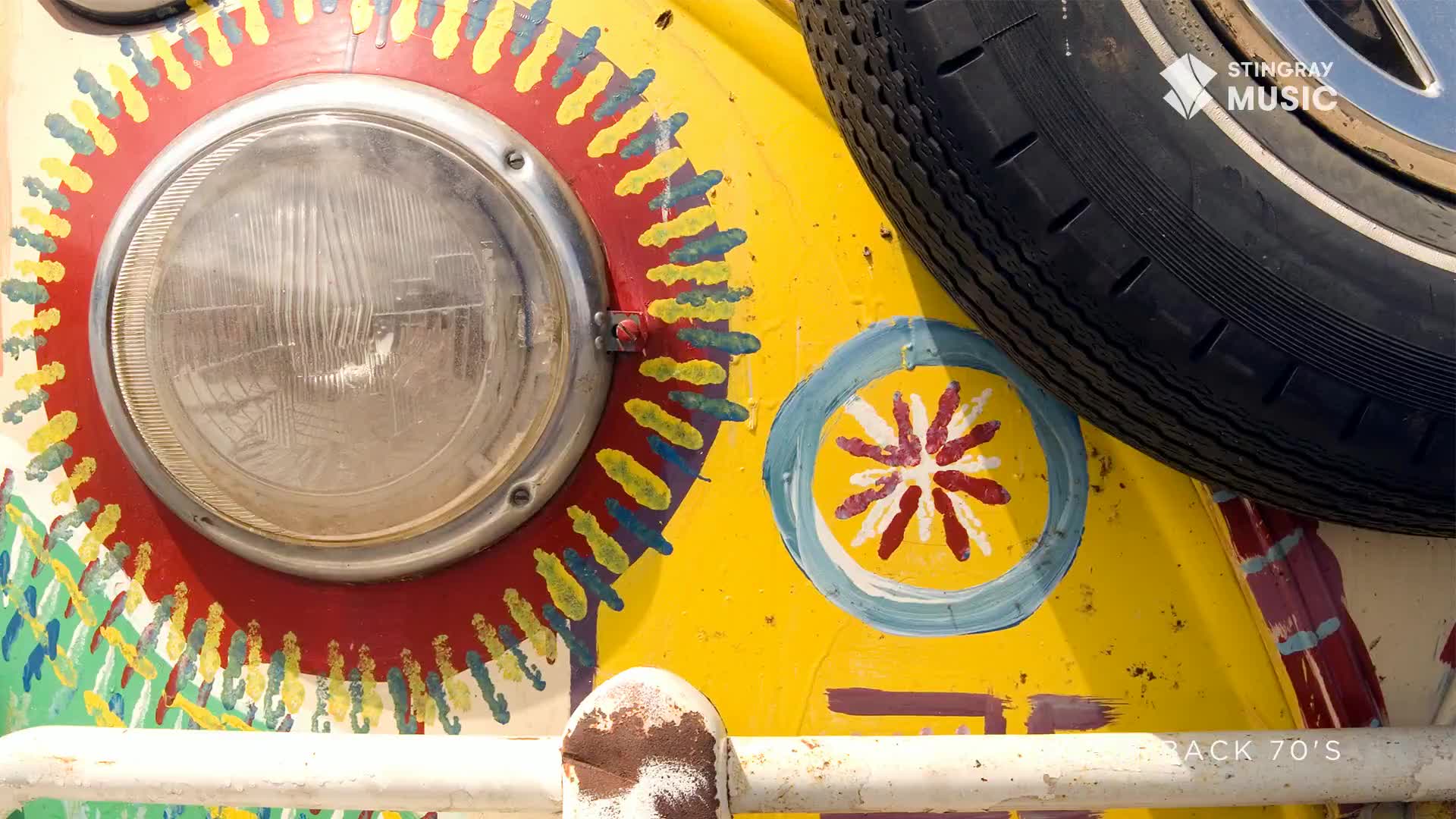 A yellow vintage van, painted with a sunburst design, sits parked. The headlight gleams, reflecting the light, as the spare tire rests on top.
