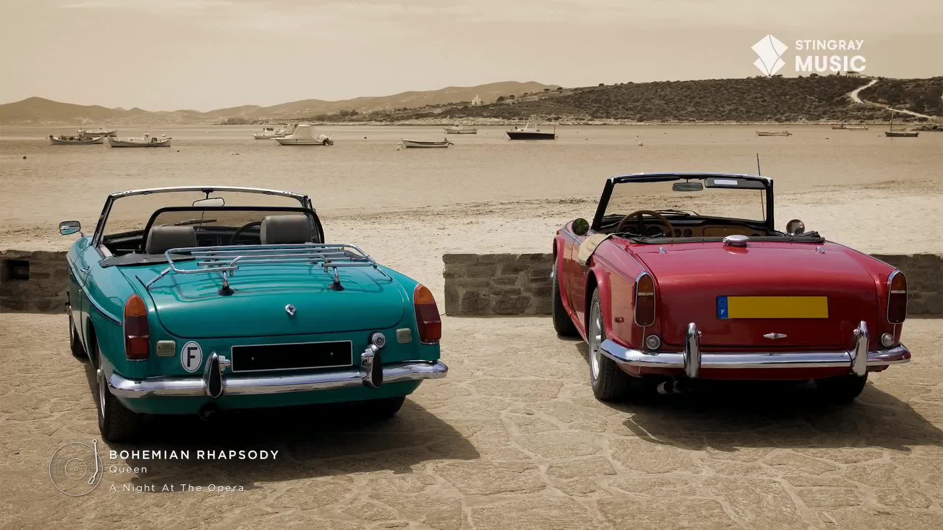 Two classic convertibles, a teal MGB and a red Triumph TR6, are parked on a cobblestone path overlooking a calm bay dotted with small boats. The scene evokes a sunny, carefree vibe, reminiscent of a 70s road trip through the Canadian countryside.
