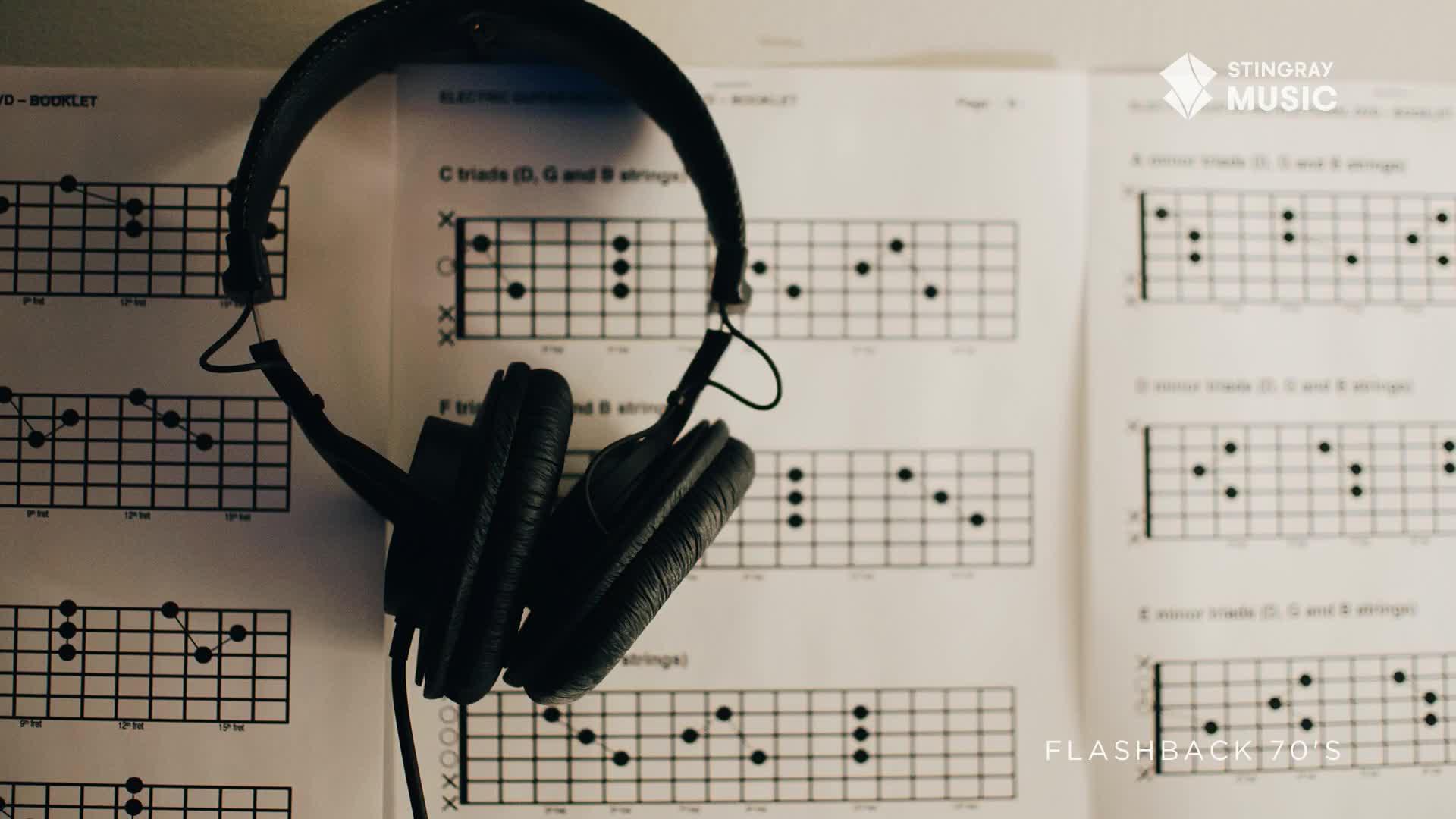 A pair of black headphones hangs over guitar chord diagrams. The papers show how to play chords, hinting at a 70s rock vibe.