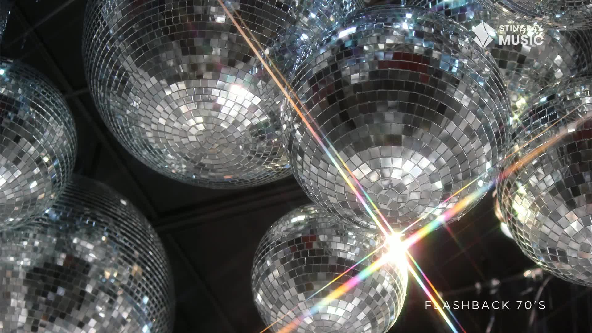 A cluster of mirrored disco balls hangs overhead, their facets catching the light in a dazzling display. Rainbow flares streak across the scene, hinting at a vibrant 70s flashback.