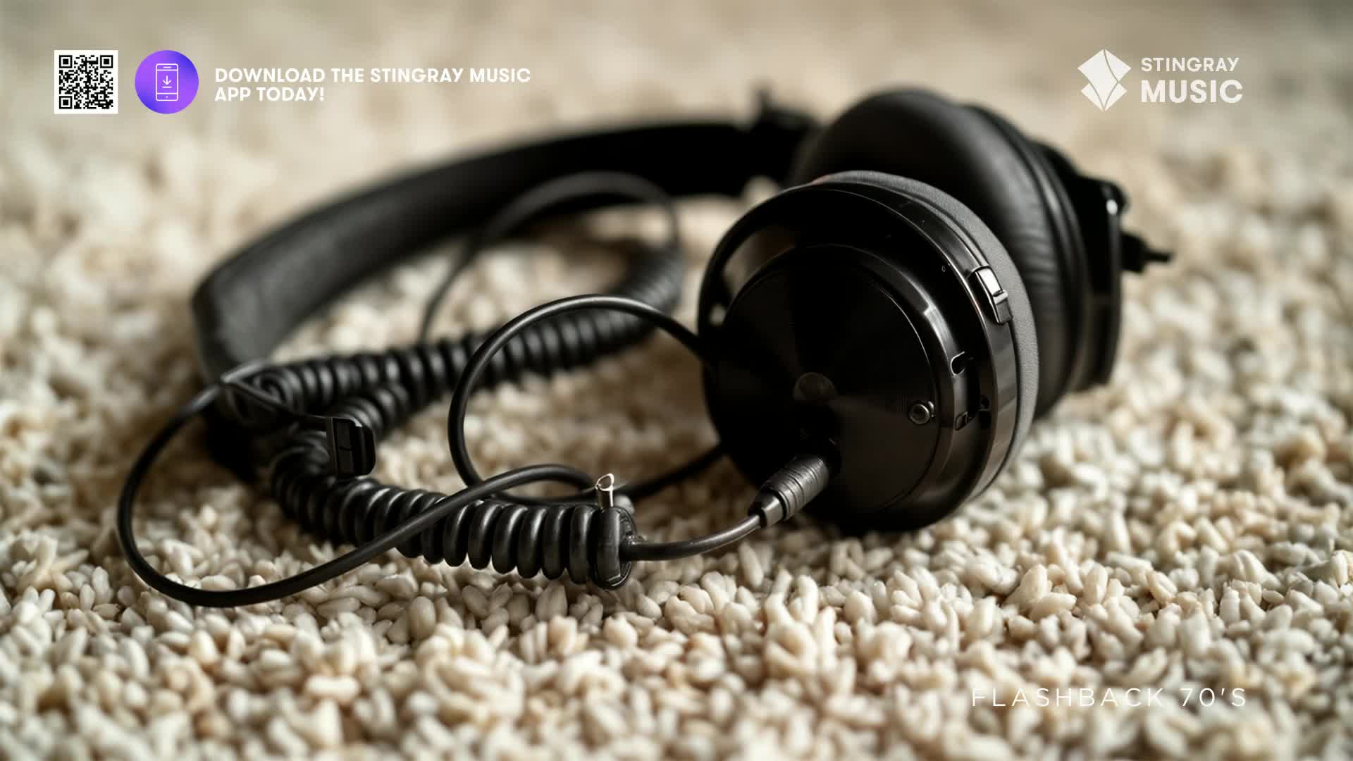 A pair of black, over-ear headphones rests on a textured carpet. The coiled cord snakes out from the earpiece, hinting at a 70s flashback.