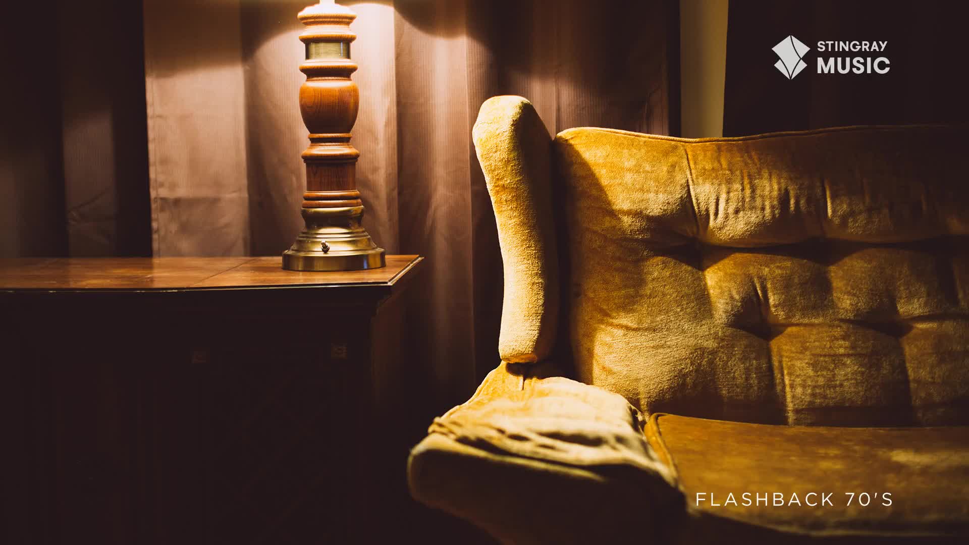 The plush, mustard-yellow velvet of a 70s armchair catches the warm glow from a wooden lamp on a side table. A faint shimmer of light plays across the tufted backrest, hinting at a cozy, dimly lit room.