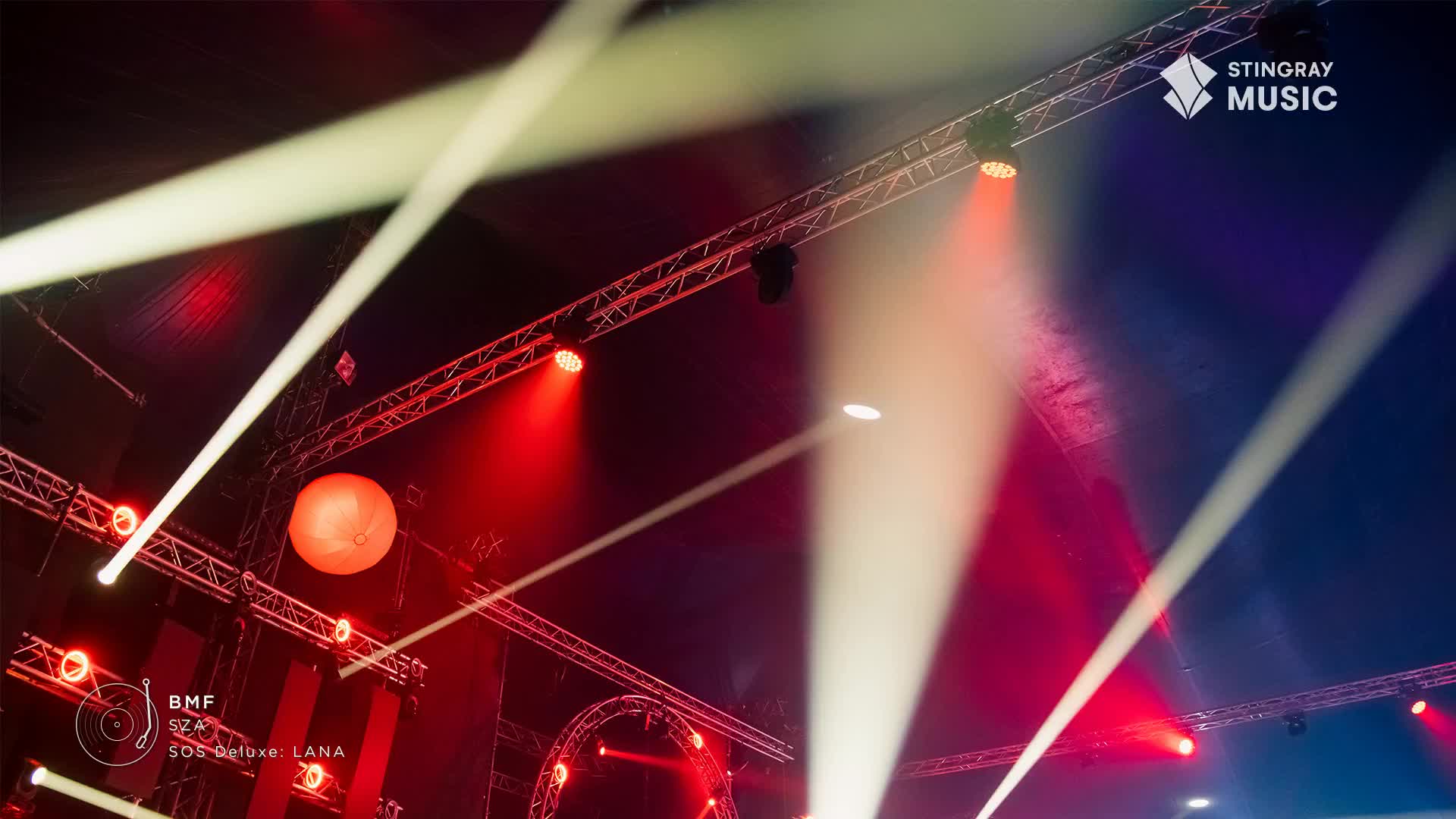 Bright white beams slice across the dark, crisscrossing the stage lights that are focused in crimson. The Stingray Music logo is visible, suggesting a concert in Canada.
