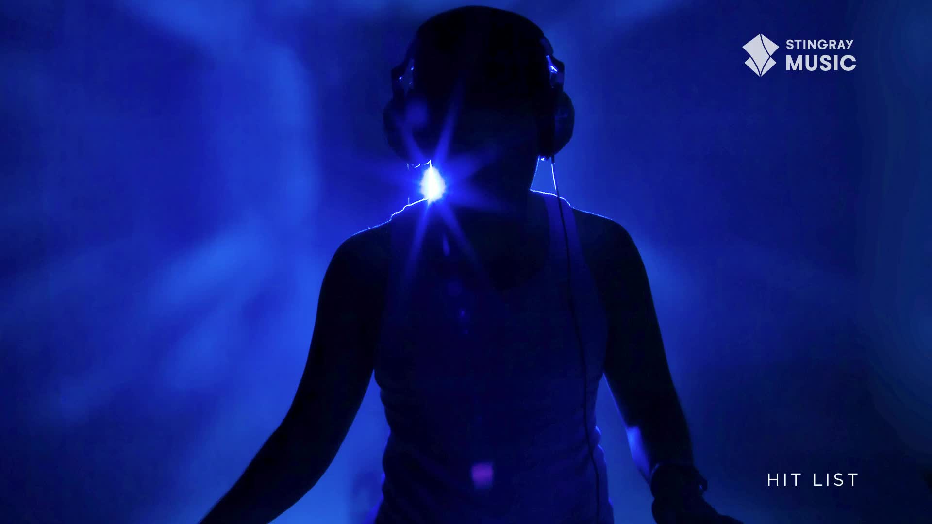 A person wearing headphones stands in a blue-lit space, their silhouette framed by the Stingray Hit List logo. The bright light behind them creates a halo effect.
