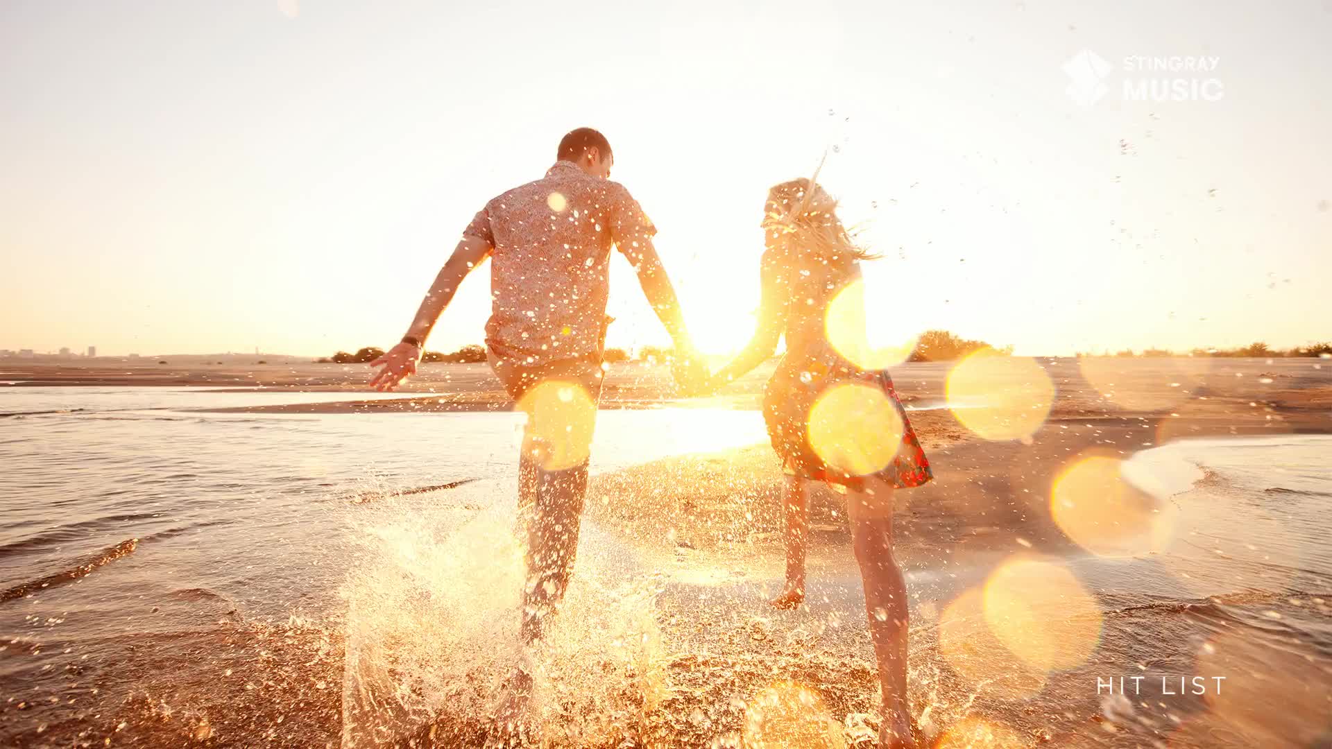 A couple, hand-in-hand, runs through the shallow water, kicking up sprays of water. The warm light of the setting sun bathes them as they move toward the shore, a scene that feels like it could be on a playlist from Stingray Hit List in Canada.
