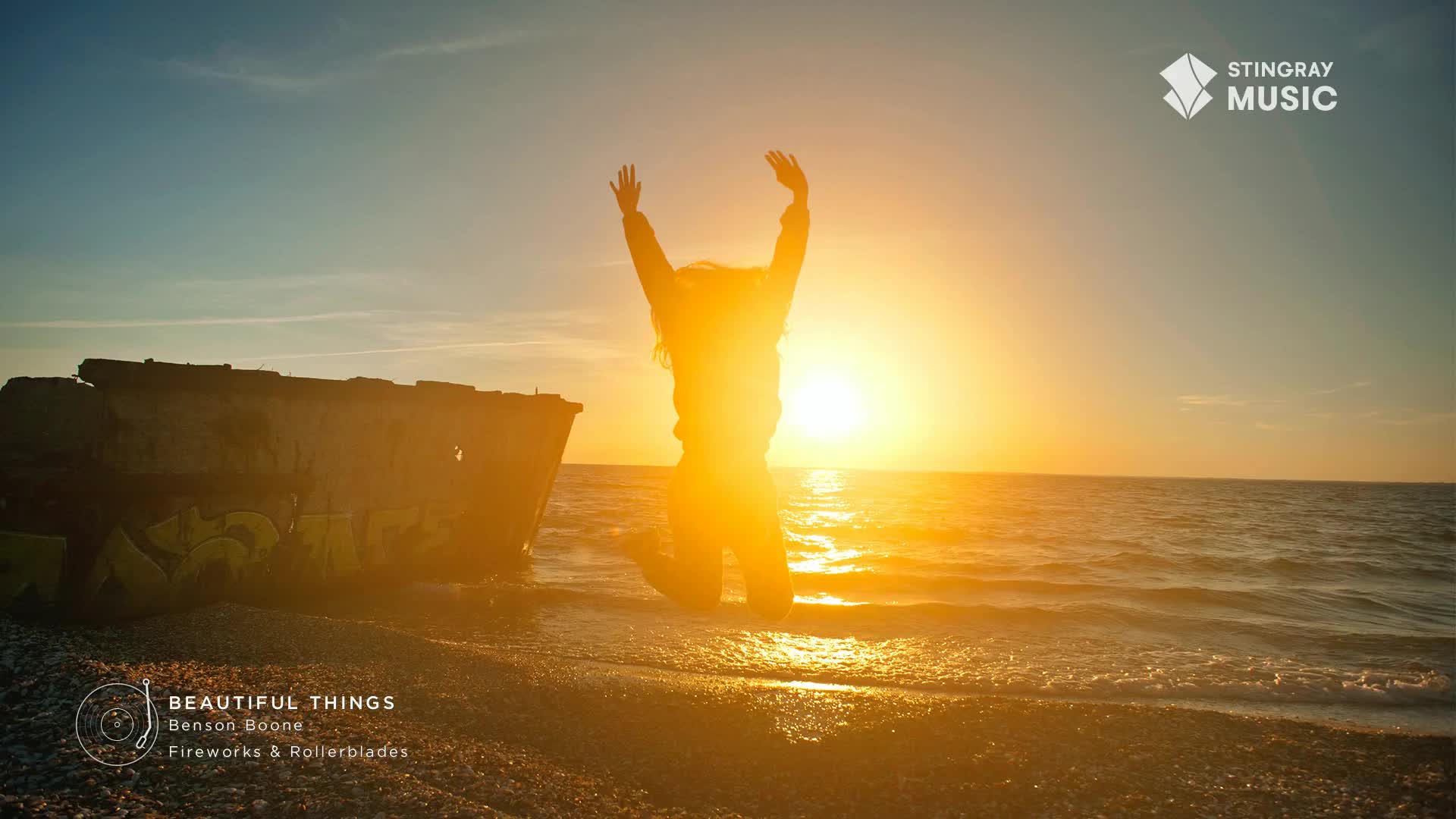 A person is mid-jump on the shoreline, arms raised in the air as the sun sets over the water. The Stingray Music logo is in the corner, and it looks like a beautiful day in Canada.
