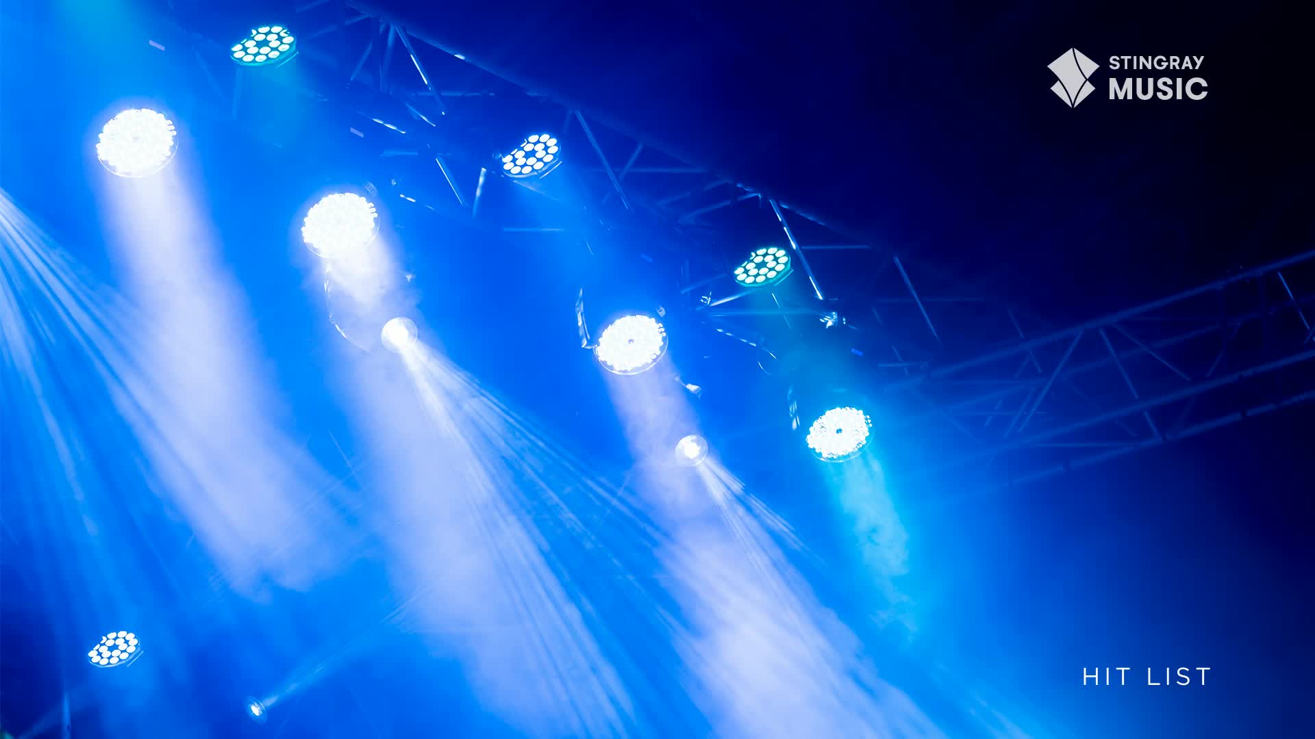 Bright blue spotlights pierce the darkness, their beams cutting through the air. Above, the metal framework of the stage supports the lights, with the Stingray Hit List logo visible in the corner.
