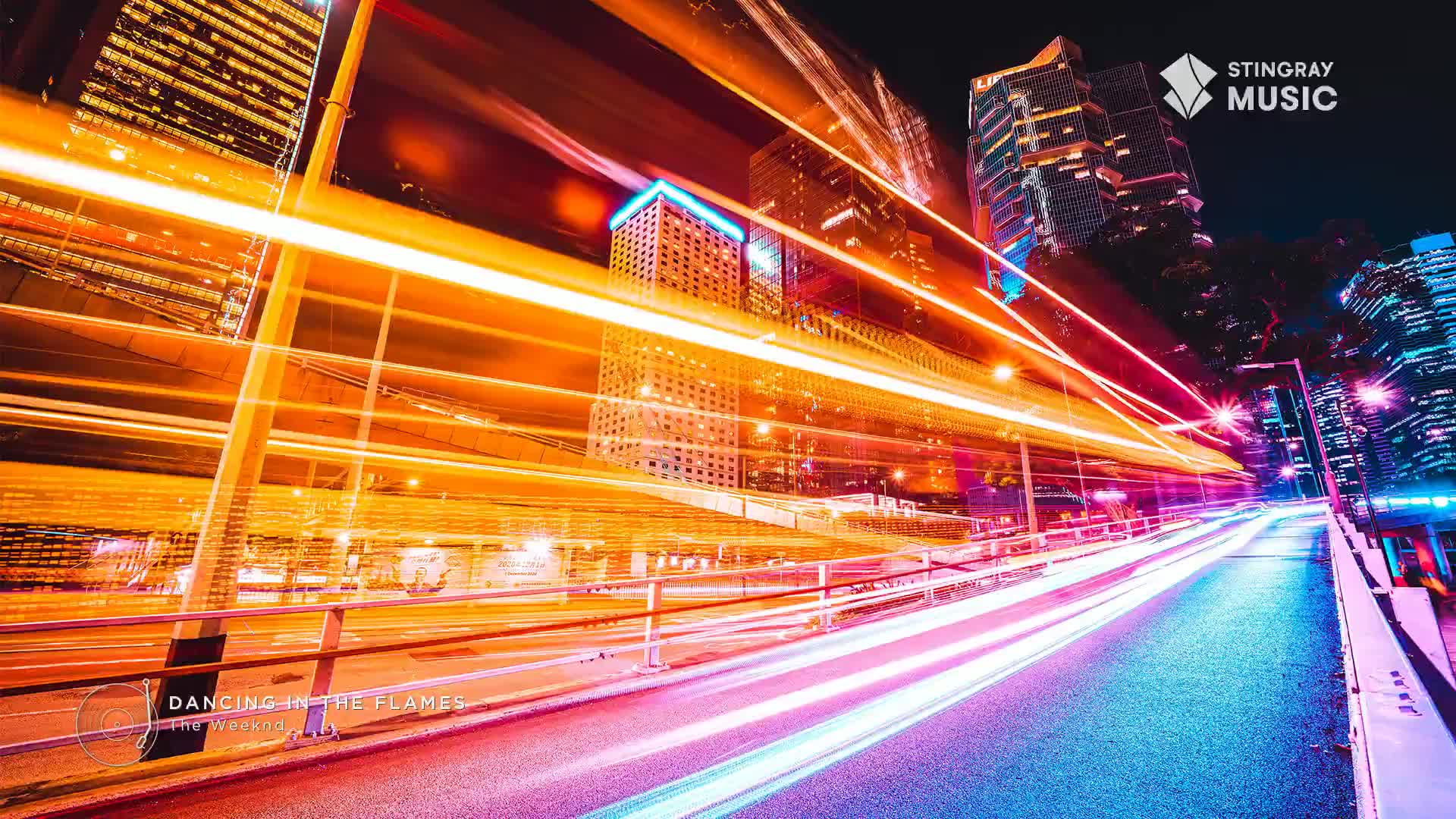 Streaks of light, likely from vehicles, race down a city street, creating long, orange and white trails against the night sky. The Stingray Music logo and "Dancing in the Flames" title card, both from Canada, are visible in the frame.
