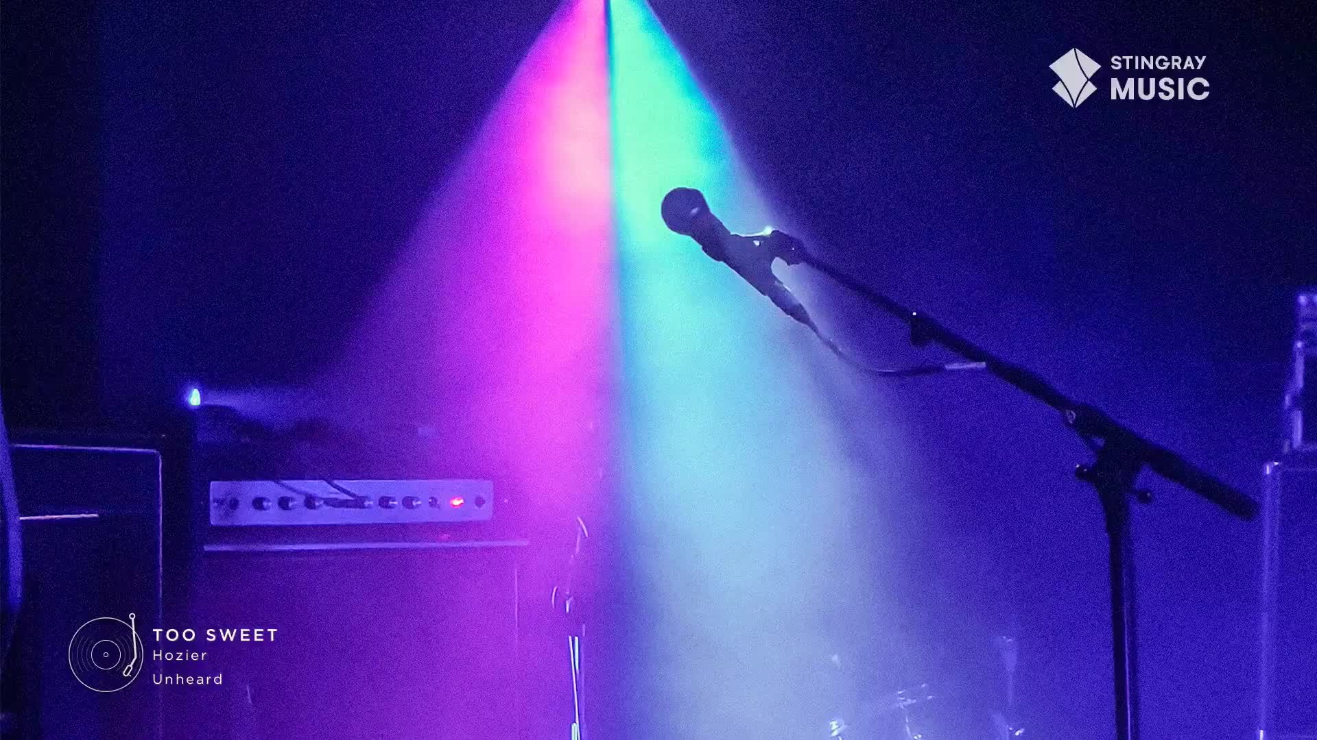 A microphone stands ready under a spotlight, its head catching the purple and green beams. The stage is set for Hozier, a Stingray Hit List performance.
