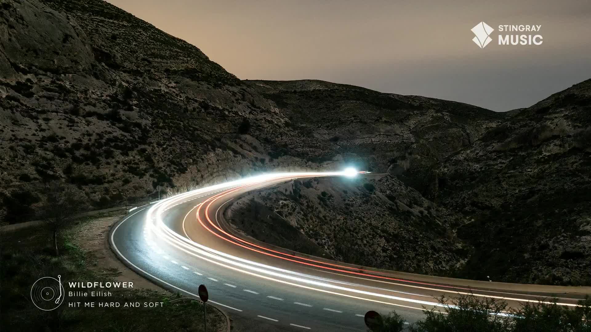 Headlights streak along a winding road, leaving glowing trails against the dark mountain. The Stingray Music logo sits in the top right corner, and the Billie Eilish "Hit Me Hard and Soft" album art is in the lower left.
