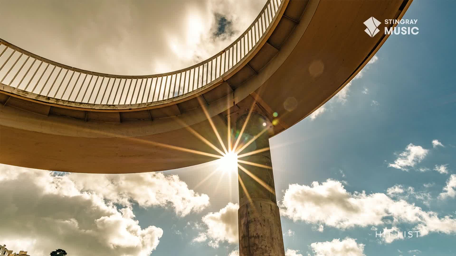 The sun bursts through the concrete support, casting a starburst of light. Above, a curved walkway with a railing arcs across the sky.
