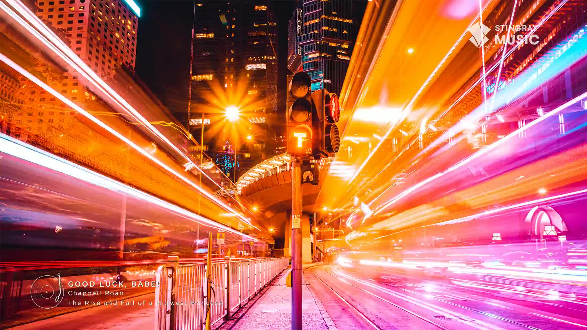 The traffic light glows orange against a backdrop of streaks, likely from passing vehicles. Buildings tower in the distance, illuminated by city lights, while the Stingray Music logo sits in the upper right.
