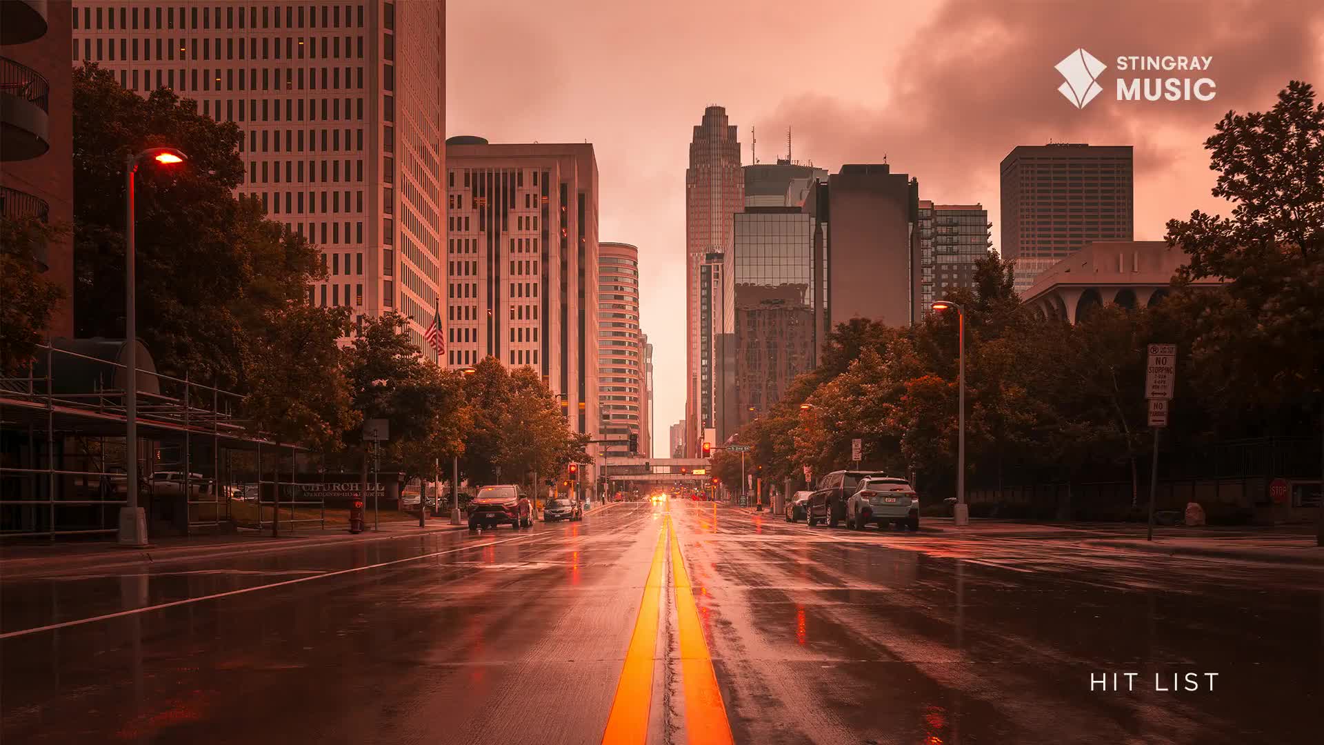 The rain-slicked street reflects the city lights, leading the eye toward the distant skyscrapers. Cars slowly navigate the wet pavement, framed by the tall buildings and trees of a Canadian city.

