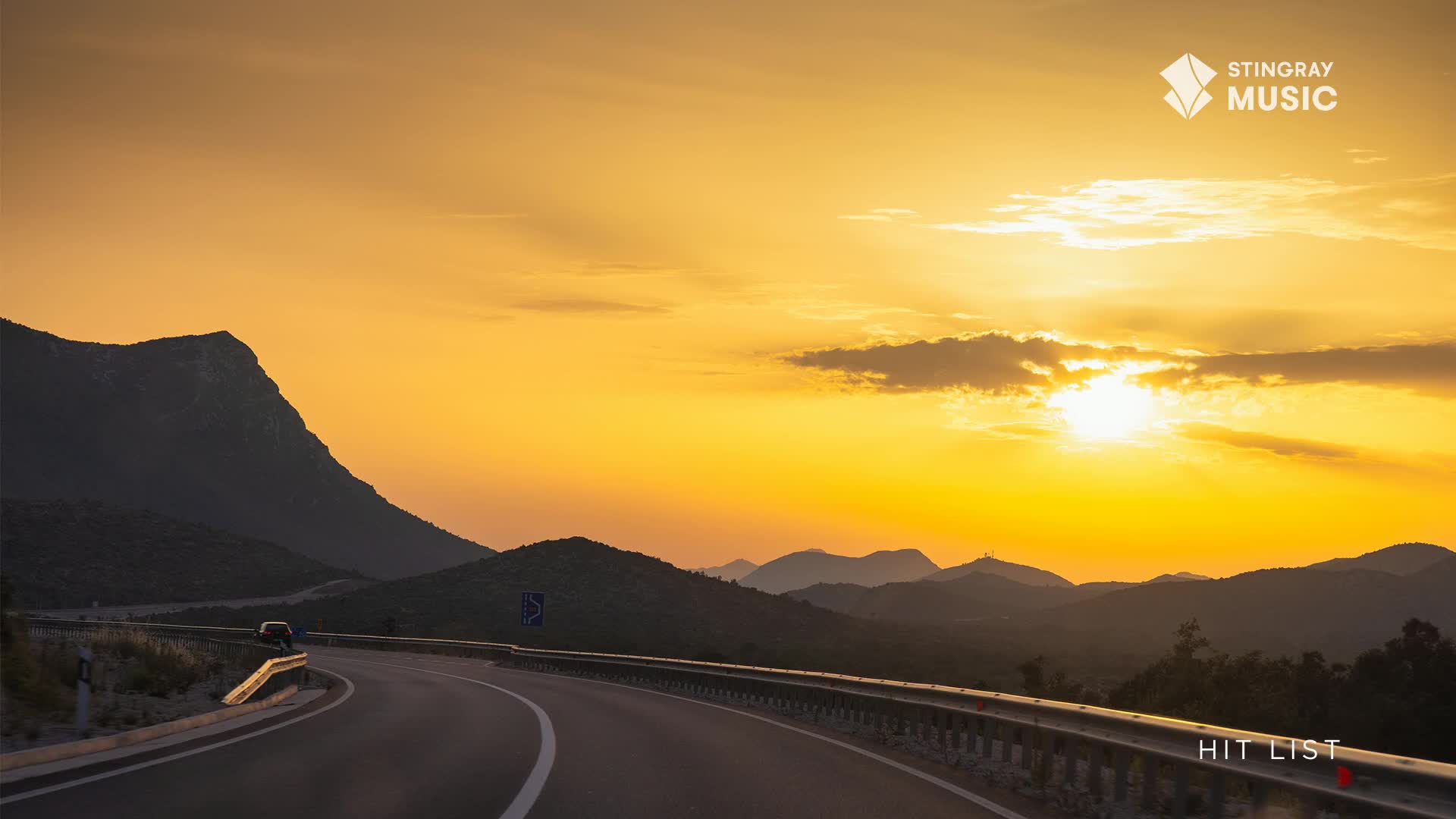 The sun is setting, painting the sky and the distant mountains in warm hues. A car is driving along a winding road, maybe headed to a Stingray Hit List show in Canada.
