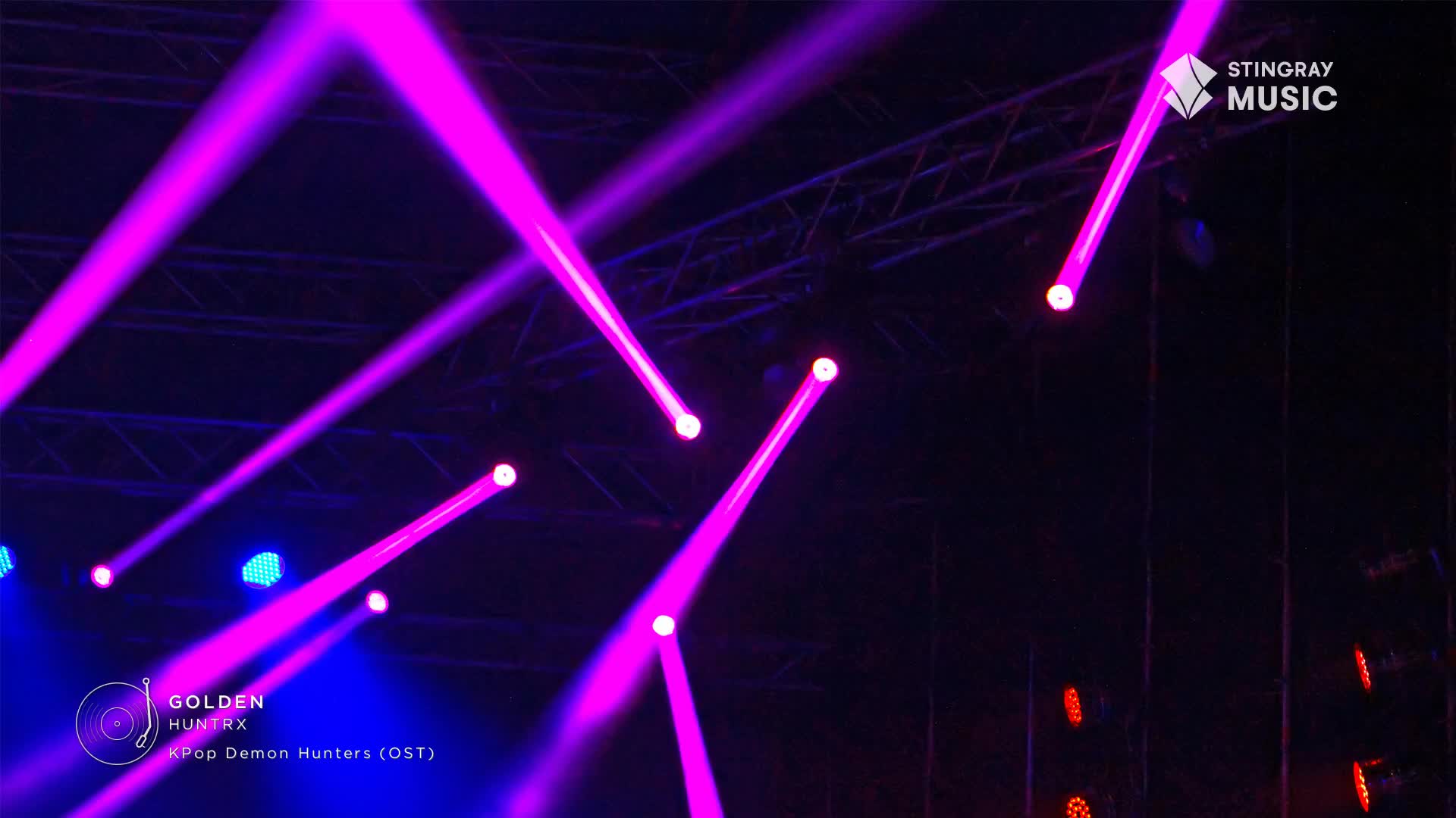 Bright purple beams of light cut across the dark stage, crisscrossing above the Stingray Music logo. The lights, part of a performance in Canada, illuminate the metal framework overhead.
