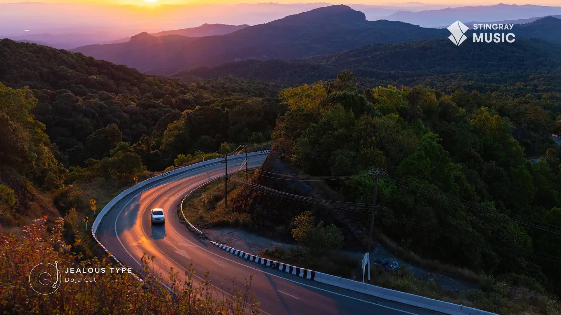 A white car cruises along a winding road, bathed in the warm glow of the sunset. The Stingray Music logo sits in the corner, as the song "Jealous Type" by Doja Cat plays on the radio.
