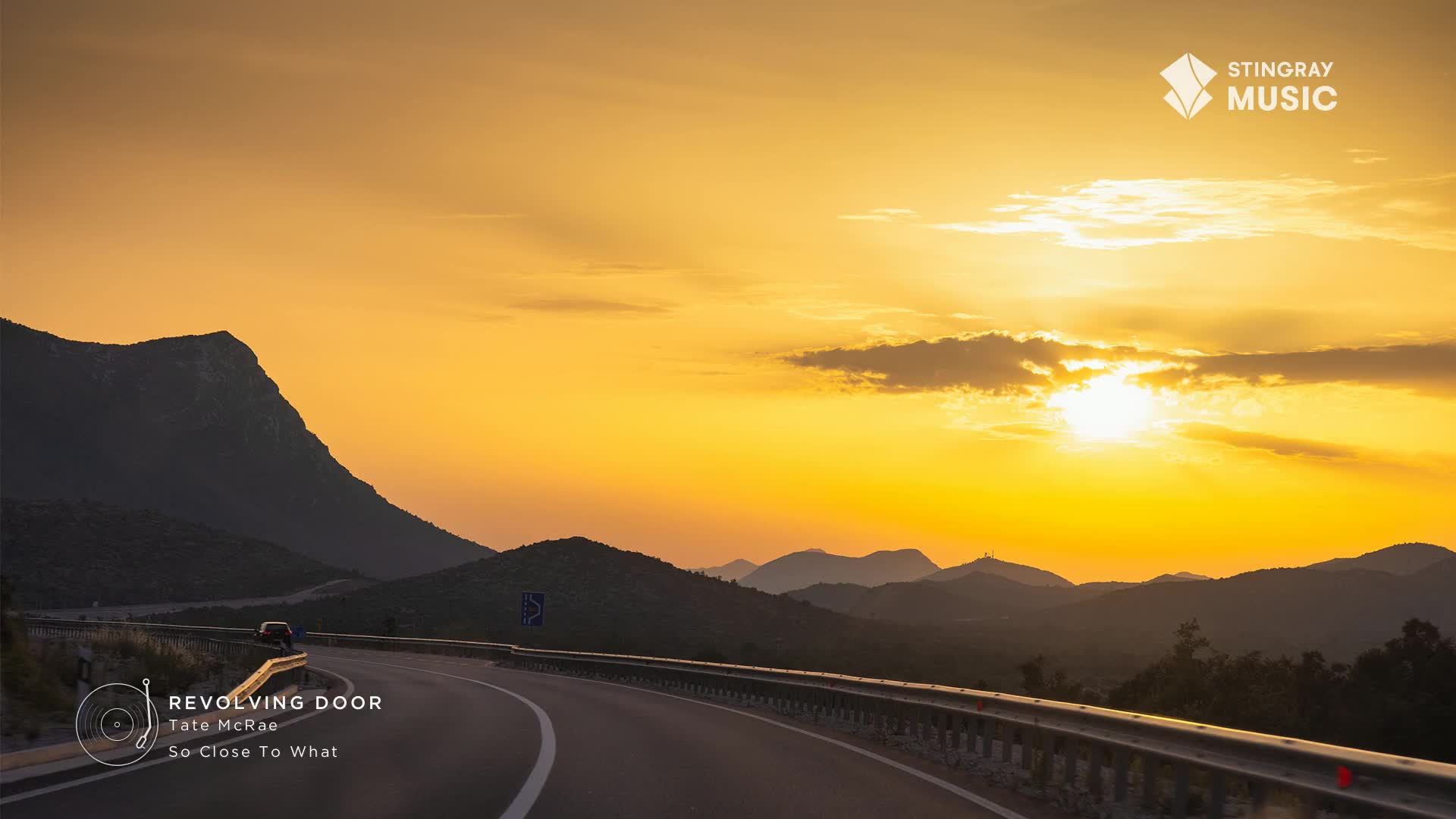 The sun is setting over a winding road, casting a warm glow across the sky. A car is heading down the road, likely towards another destination, as the Stingray Hit List plays in Canada.
