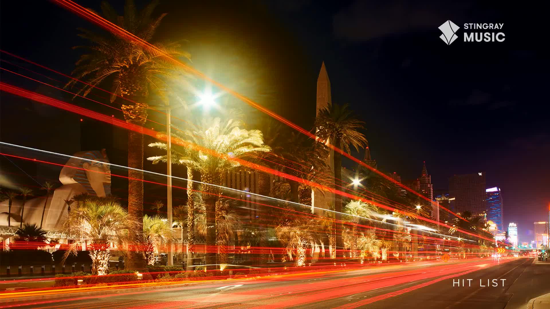 Red streaks of light, from passing vehicles, cut across the night sky above the palm trees. The Stingray Hit List is playing in Canada, and the city's bright lights are visible in the distance.
