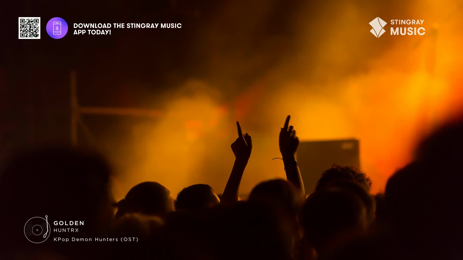 The crowd is packed, their arms raised, fingers pointing towards the stage. Warm stage lights illuminate the air, creating a hazy, golden glow.
