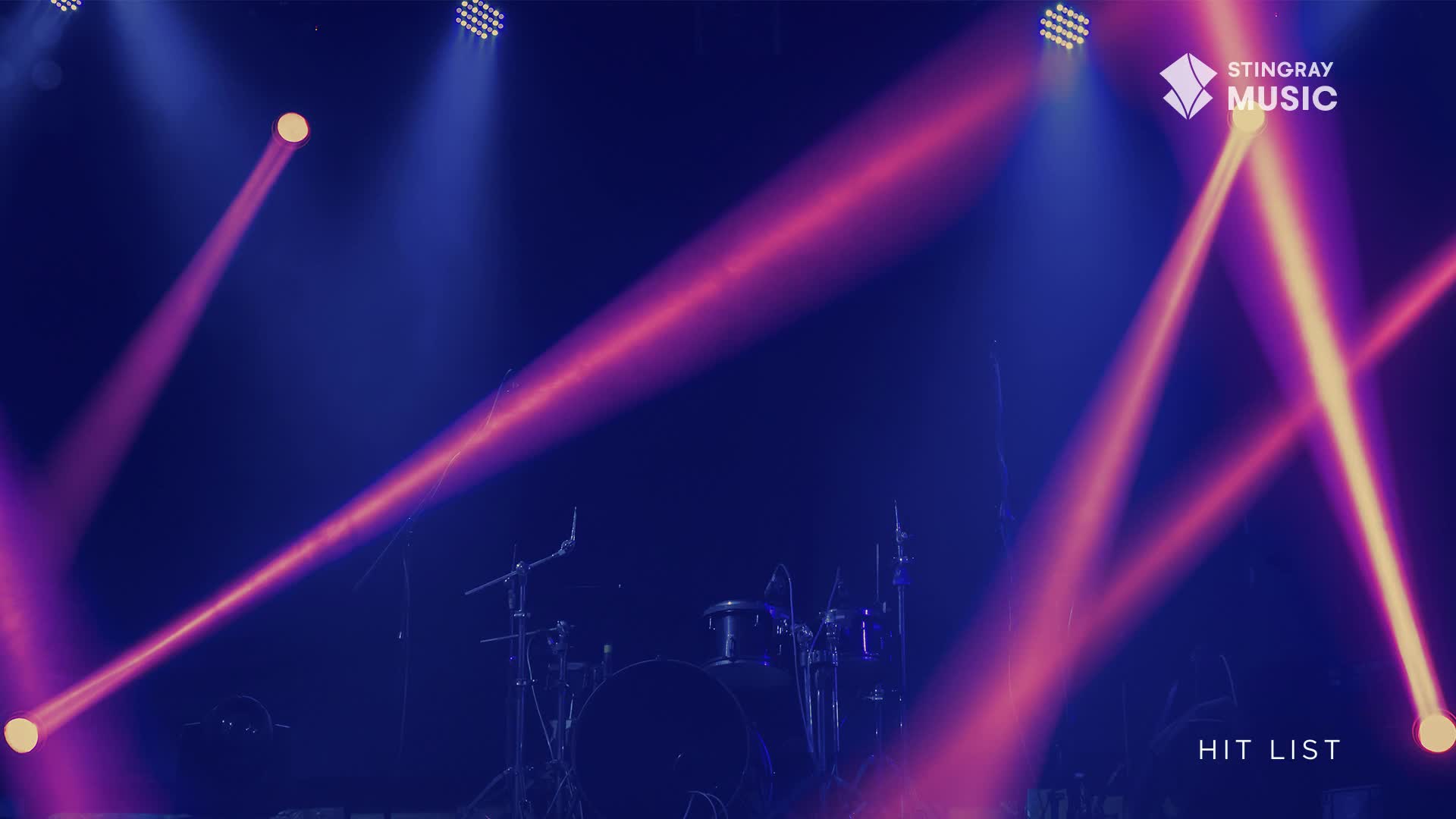 Pink and yellow beams slice through the dark, illuminating the drum kit and mics. The Stingray Hit List logo is visible, and the stage is set for a show.
