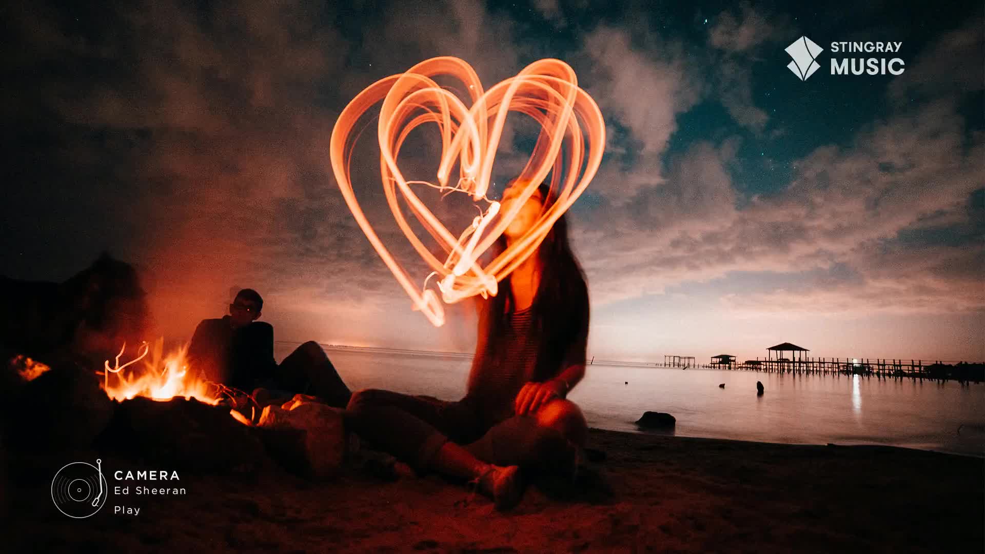 A person is drawing a glowing heart in the air with a light source, the streaks of light illuminating the dark sky. A campfire burns on the beach, casting an orange glow on the scene as the Stingray Music logo appears.
A person is drawing a glowing heart in the air with a light source, the streaks of light illuminating the dark sky. A campfire burns on the beach, casting an orange glow on the scene as the Stingray Music logo appears.