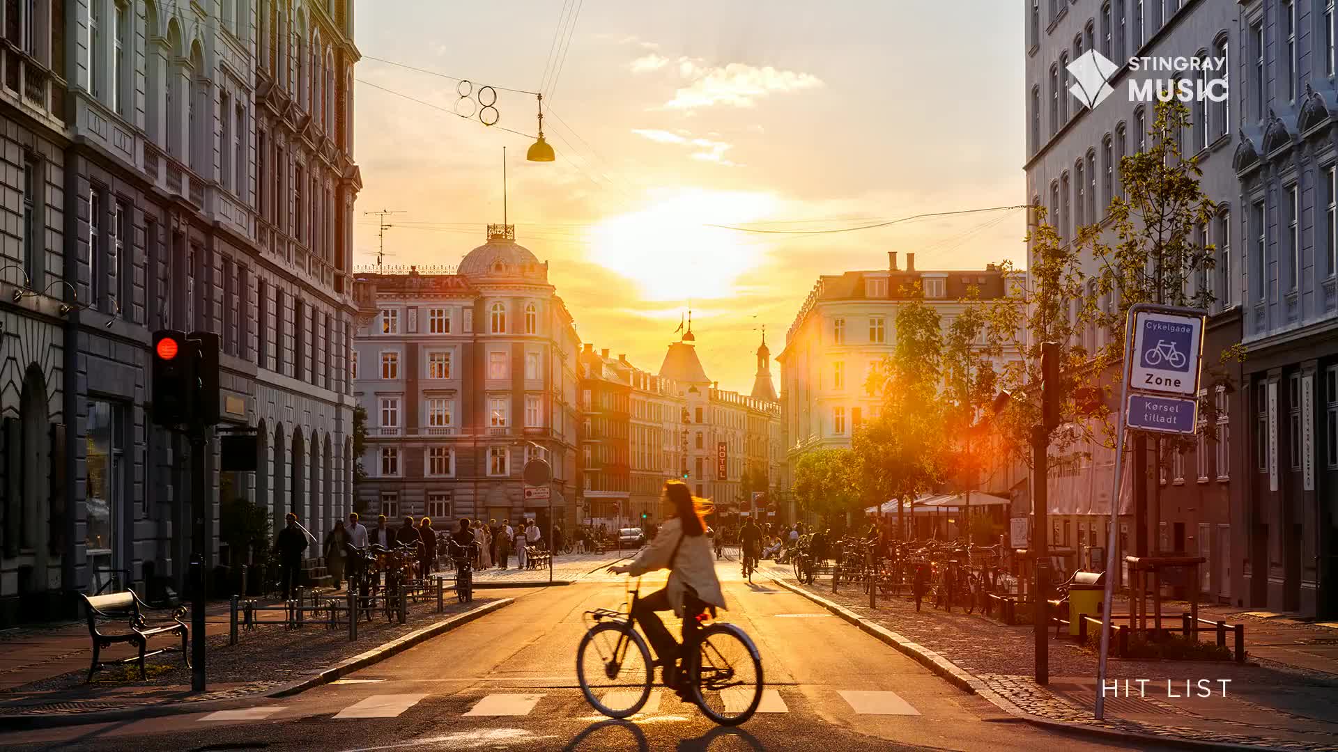 A cyclist pedals through a crosswalk, bathed in the warm glow of a setting sun. Buildings line the street, their facades catching the light as the day winds down.