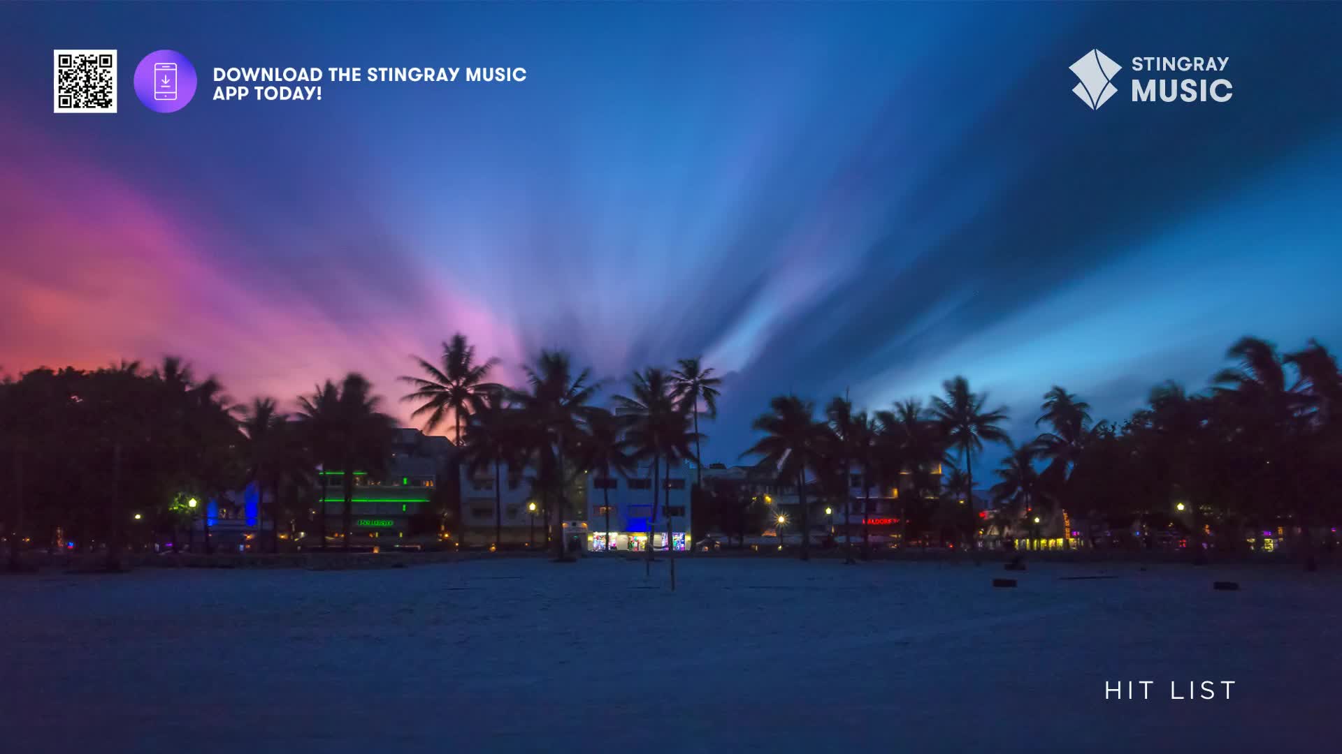 The sky bleeds purple and blue over a line of palm trees and buildings.  Lights from the town twinkle behind the silhouetted fronds.
