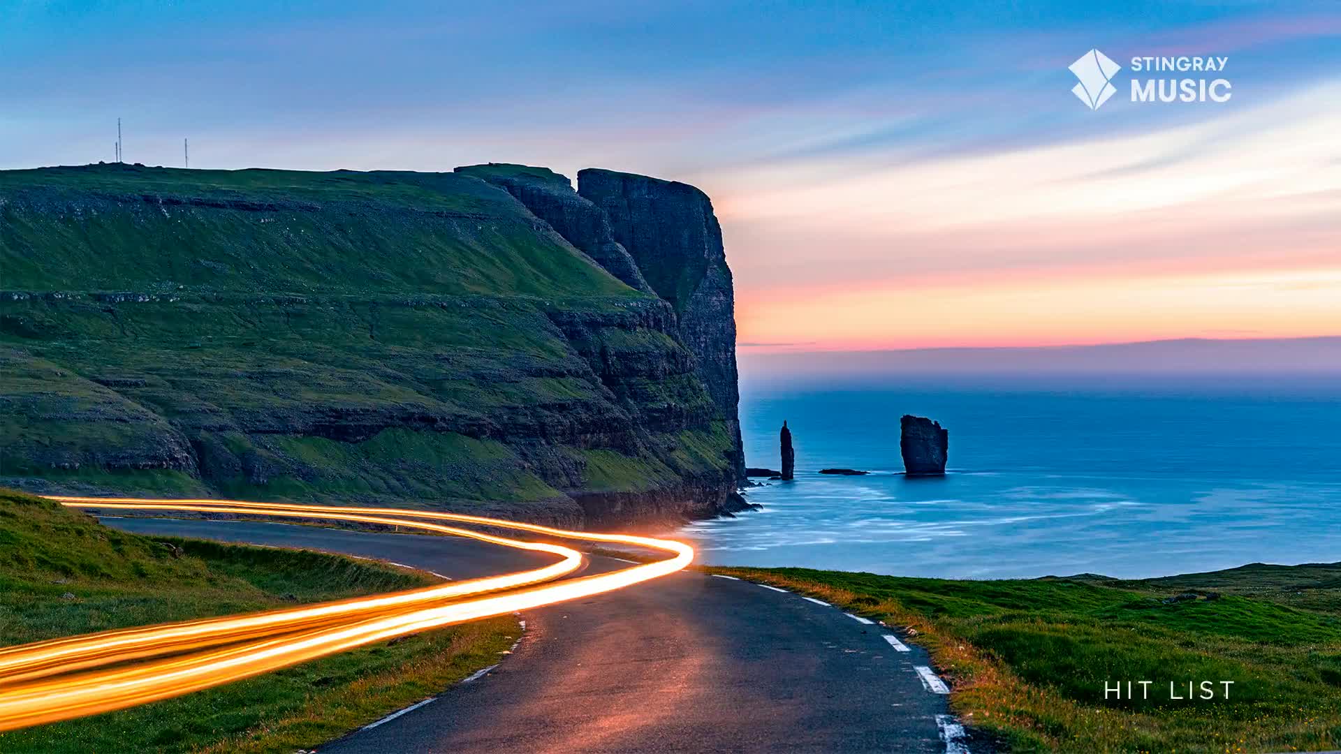 Headlights streak along the winding road, painting the asphalt with long, glowing lines. The ocean stretches out to the horizon, meeting a sky awash in soft twilight hues.