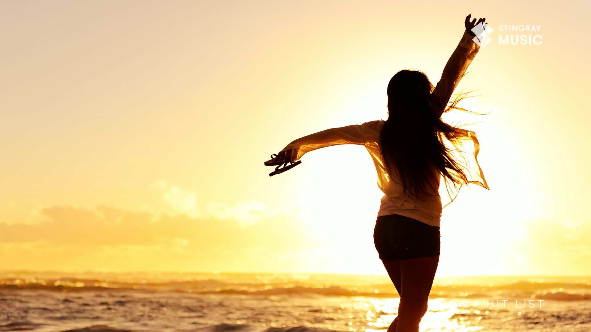 A young woman with long dark hair throws her arms wide, holding a pair of flip-flops. The setting sun casts a warm glow over the ocean and beach.