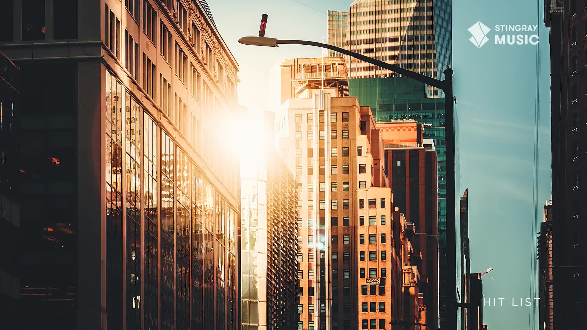 Sunlight glints off the glass facades of towering buildings as a streetlamp arcs overhead. The Stingray Hit List logo appears in the clear sky.