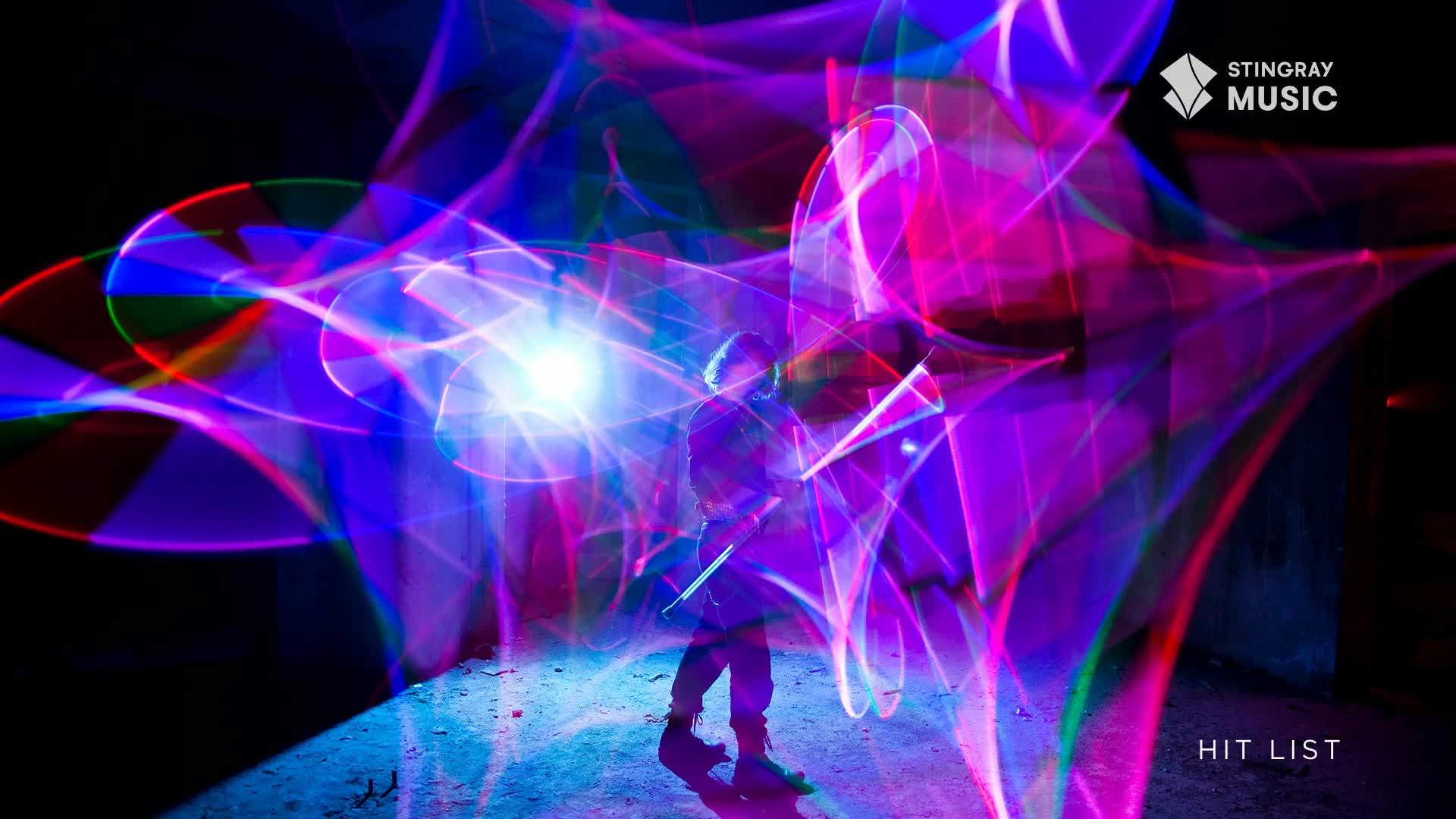 A person stands in the middle of a swirling vortex of bright pink and blue light trails, like a live-action Stingray Hit List segment. The light painting creates a dynamic, almost electric atmosphere that engulfs the figure.