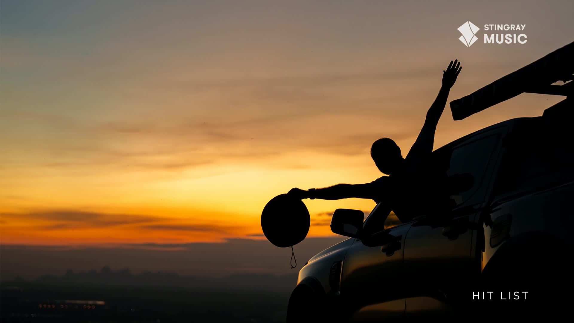 The sun dips below the horizon, painting the sky in shades of orange and gold. A person leans out of a car window, their silhouette stark against the vibrant backdrop, a black hat dangling from their hand.