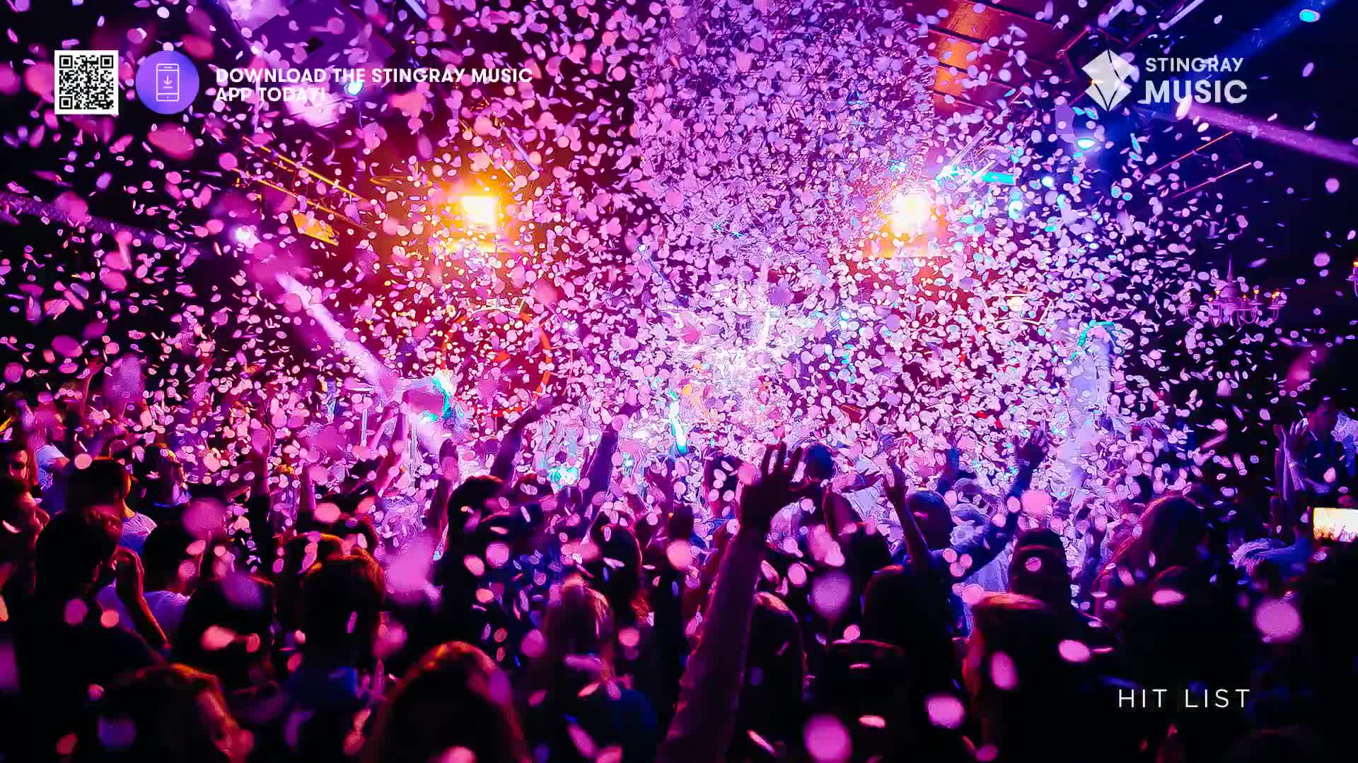 Confetti rains down on a packed crowd, their hands reaching up. The air is thick with pink and purple paper, illuminated by stage lights.