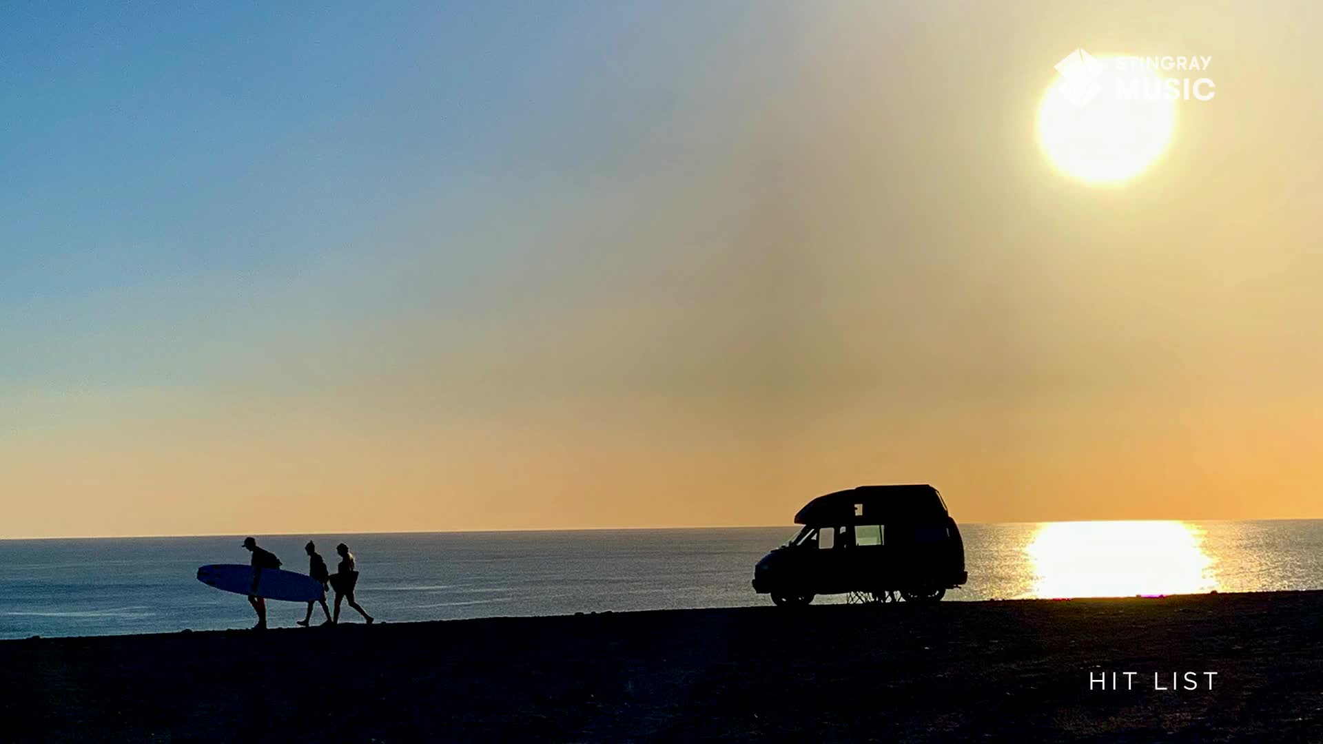 Three figures walk along the coast, one carrying a surfboard. A camper van sits parked nearby as the sun sets over the water.