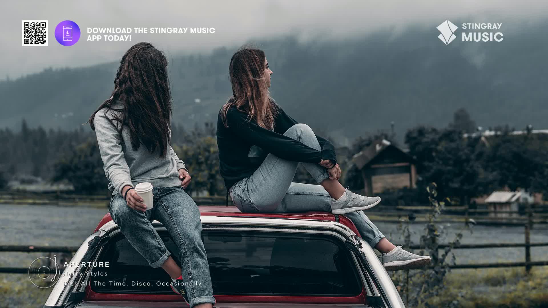 Two women sit on the back of a red truck, overlooking a misty Canadian landscape. The woman on the left holds a coffee cup, while the one on the right rests her chin on her knees.