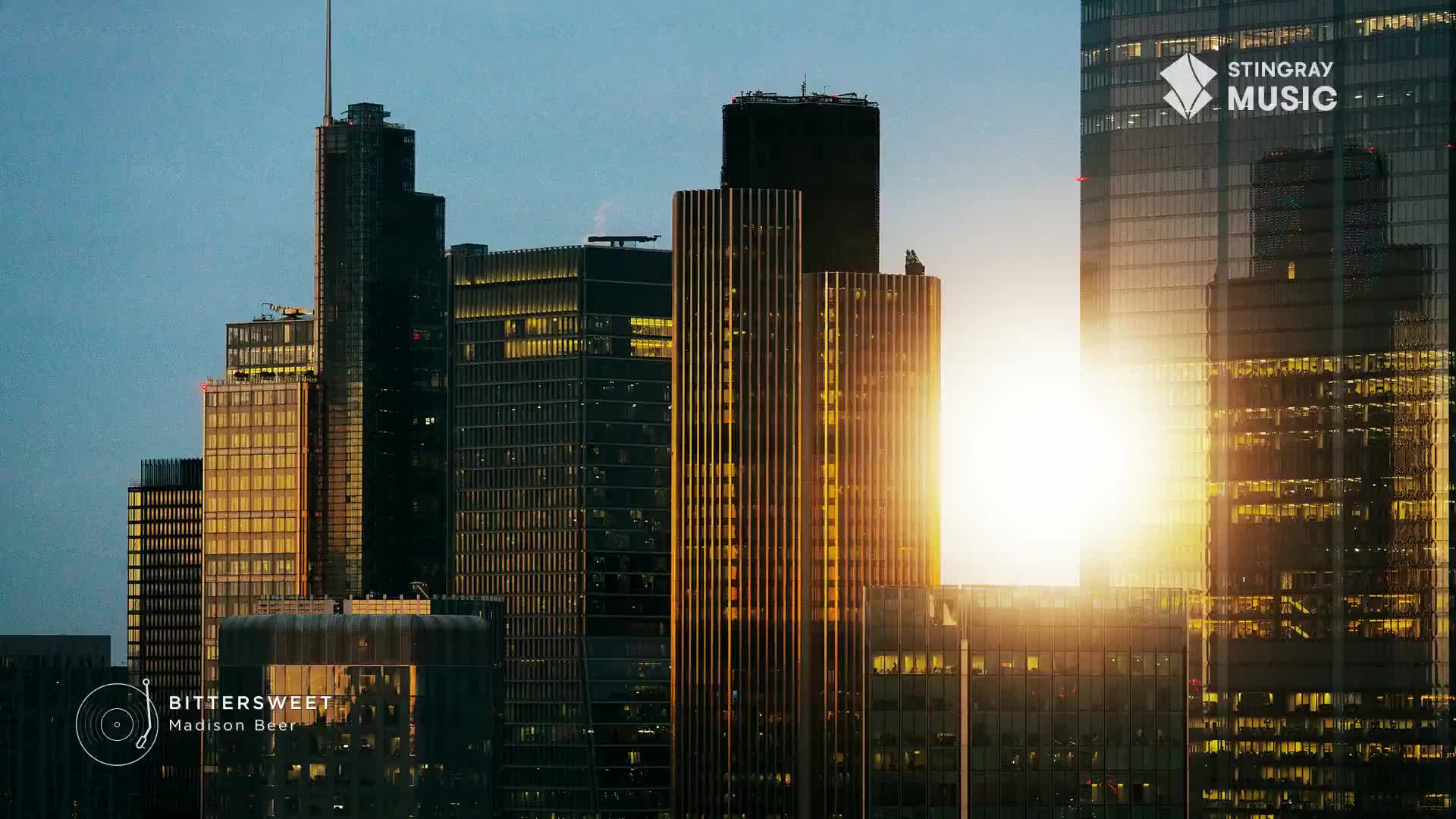 The sun is setting behind a cluster of tall buildings in downtown Toronto. Light glints off the glass facades, illuminating the city as Stingray Music's "Bittersweet" by Madison Beer plays.