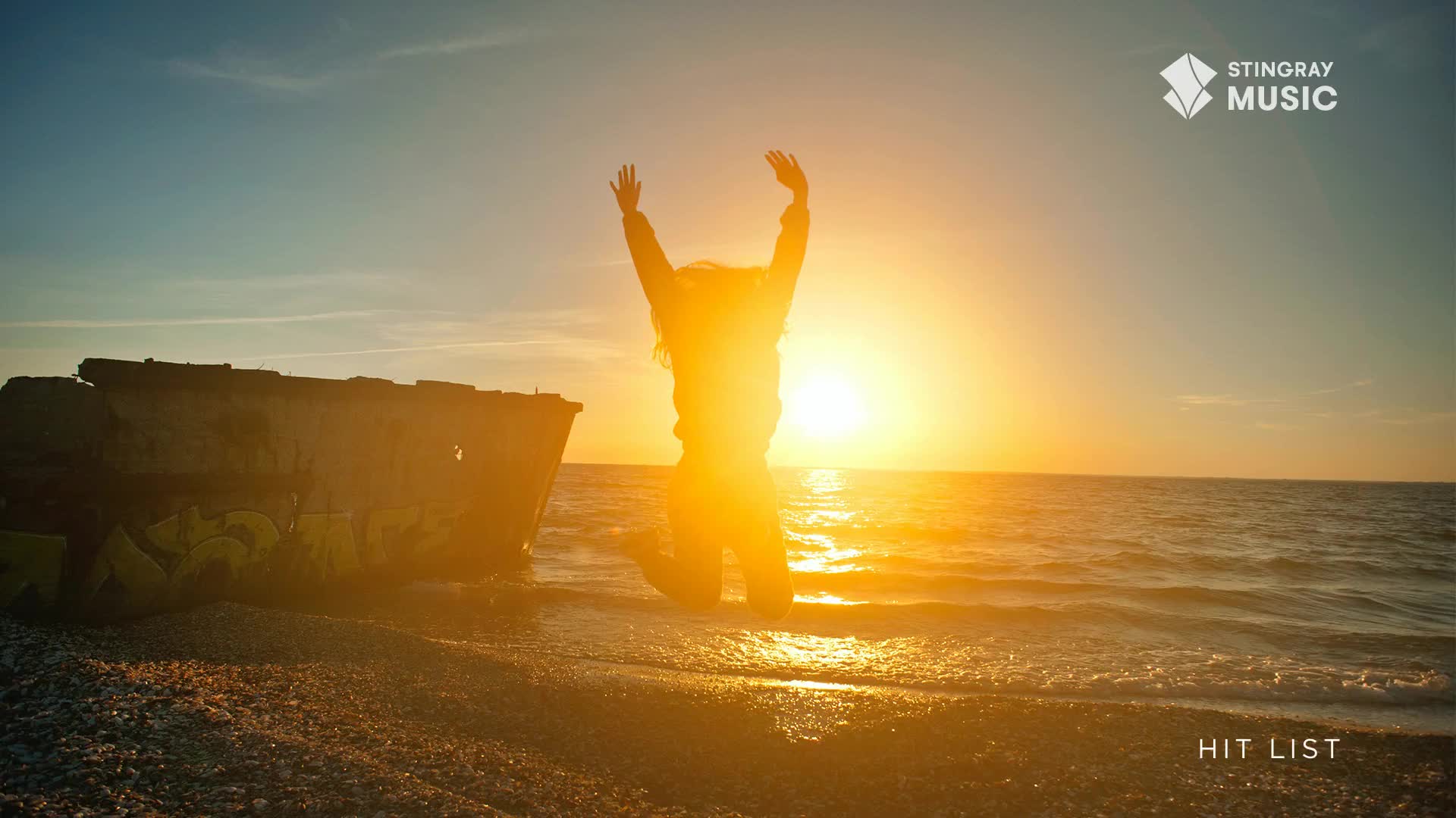 A person leaps into the air against a brilliant sunset over the water, their arms raised in triumph. Nearby, a weathered, graffiti-covered structure sits on the rocky shore, a stark contrast to the vibrant sky. A person leaps into the air against a brilliant sunset over the water, their arms raised in triumph. Nearby, a weathered, graffiti-covered structure sits on the rocky shore, a stark contrast to the vibrant sky.