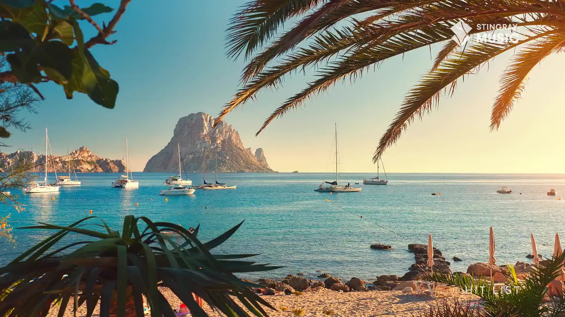 Sailboats glide across the calm, turquoise water. A massive rock formation rises from the sea in the distance.