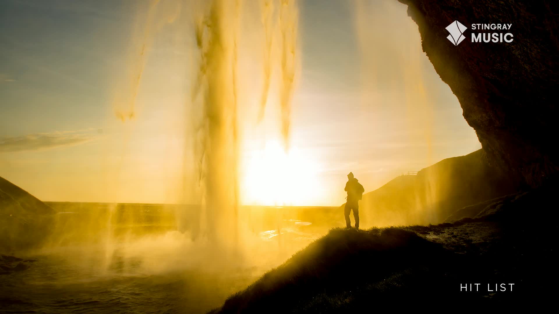 A lone figure stands on a cliff edge, silhouetted against a brilliant sunset behind a curtain of falling water. The Stingray Hit List plays as the sun dips below the horizon, casting a golden glow over the Canadian landscape.