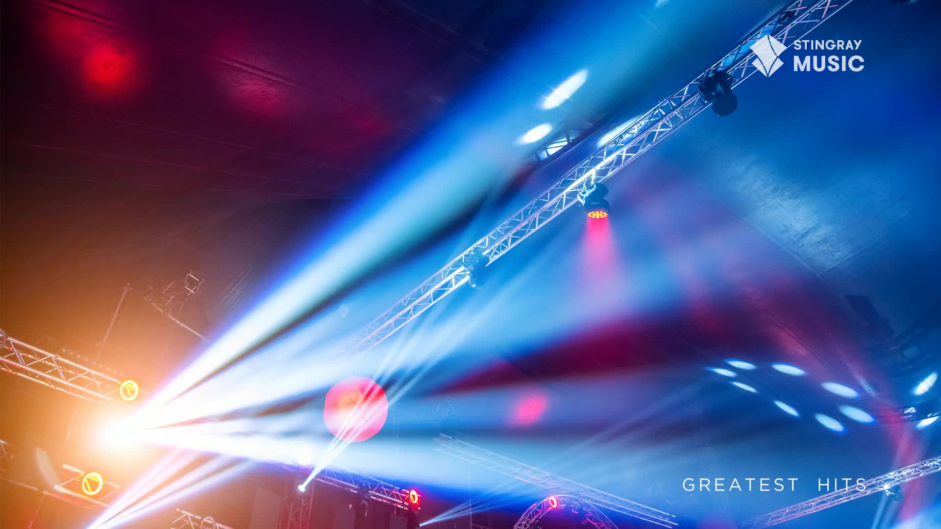 Bright beams of blue and white light slice through the air, crisscrossing above. Red and orange spotlights add bursts of color to the stage, likely for a Stingray Holiday Hits concert in Canada.
