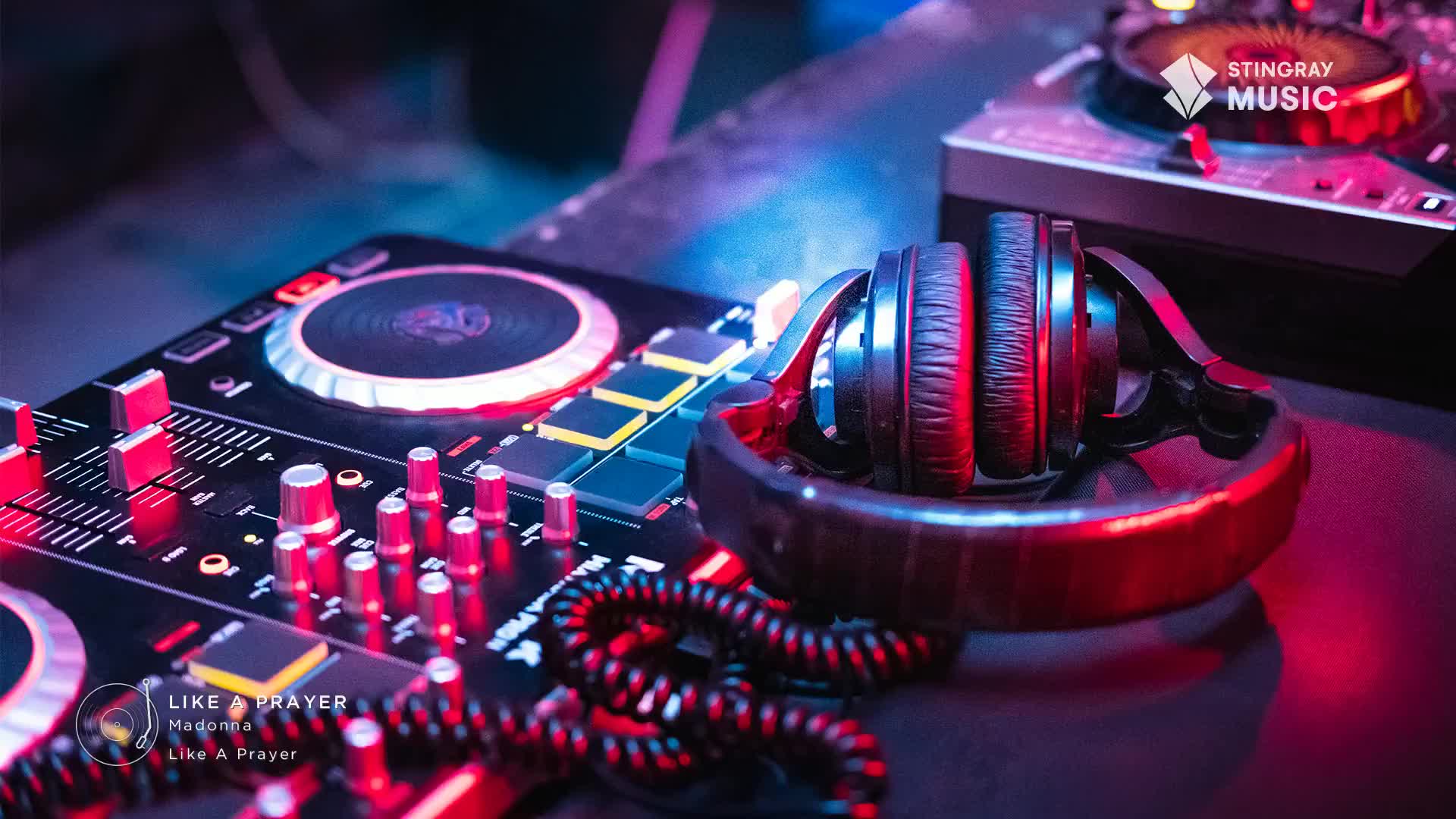 A pair of headphones rests on a stand, bathed in red and blue light, next to a DJ mixer. Knobs and buttons are illuminated, ready for the next track on Stingray Holiday Hits, maybe even Madonna's "Like a Prayer."
