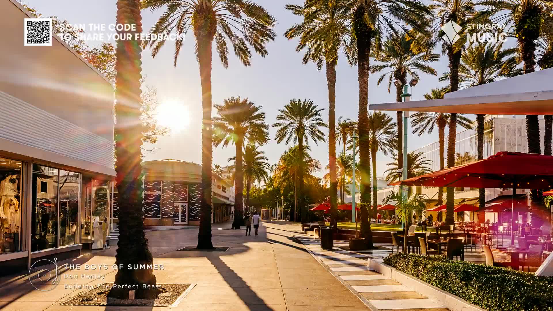 Palm trees cast long shadows across a sun-drenched walkway, and the light flares around the trunks. A few people stroll toward the back, past outdoor seating under red umbrellas, a scene that feels like a perfect summer day, the kind that might be playing on Stingray Holiday Hits in Canada.

