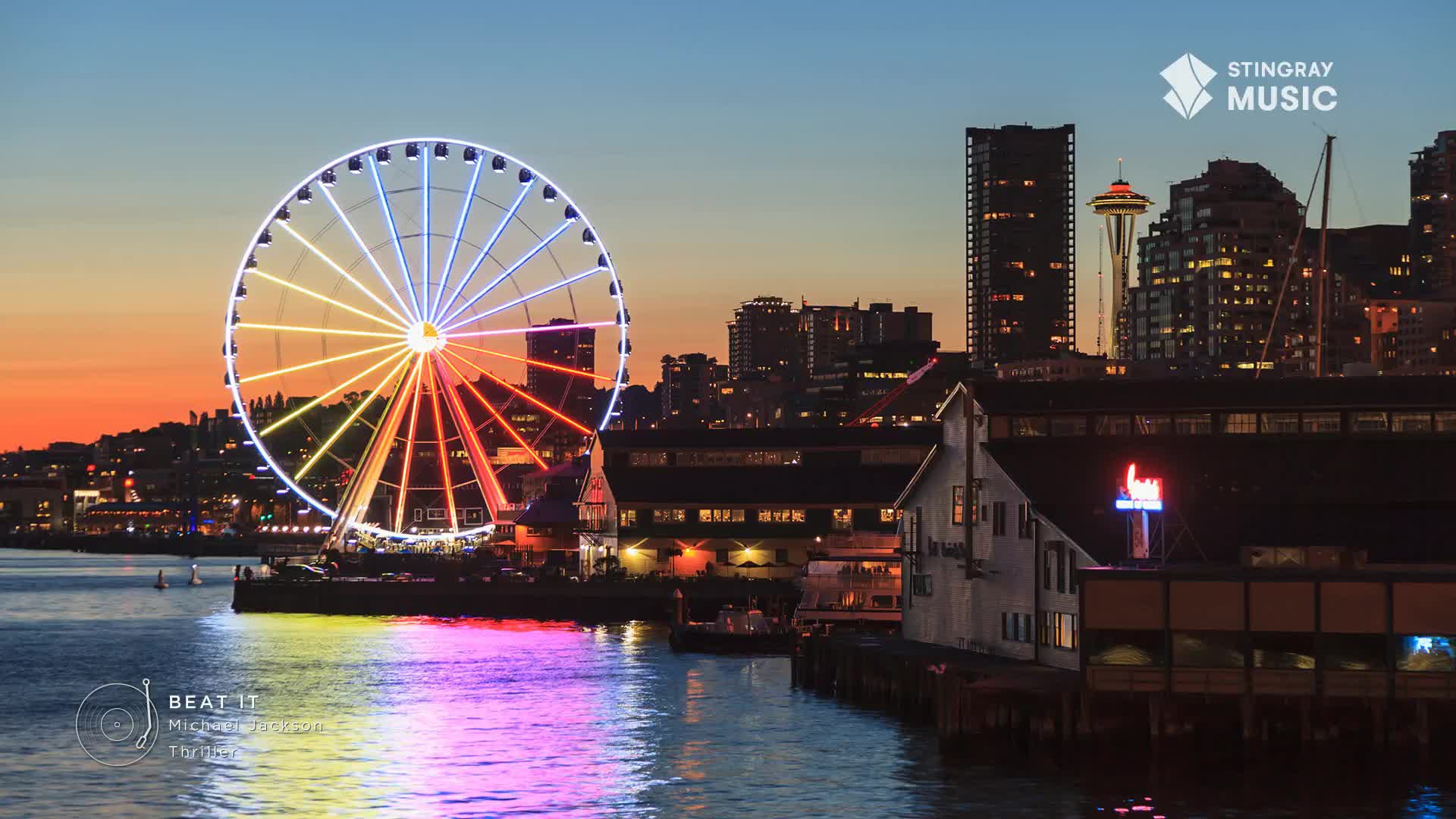 The Ferris wheel's lights shine brightly against the twilight sky, reflecting in the water below. Buildings and the Space Needle stand silhouetted in the distance, adding to the vibrant scene of Stingray Holiday Hits.
