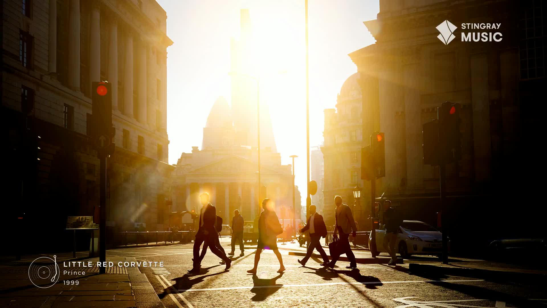 People are crossing a sun-drenched street, their silhouettes cast long by the low angle of the light. The Stingray Holiday Hits logo and a "Little Red Corvette" album graphic are in the corner, suggesting a music video or promotional image from Canada.
