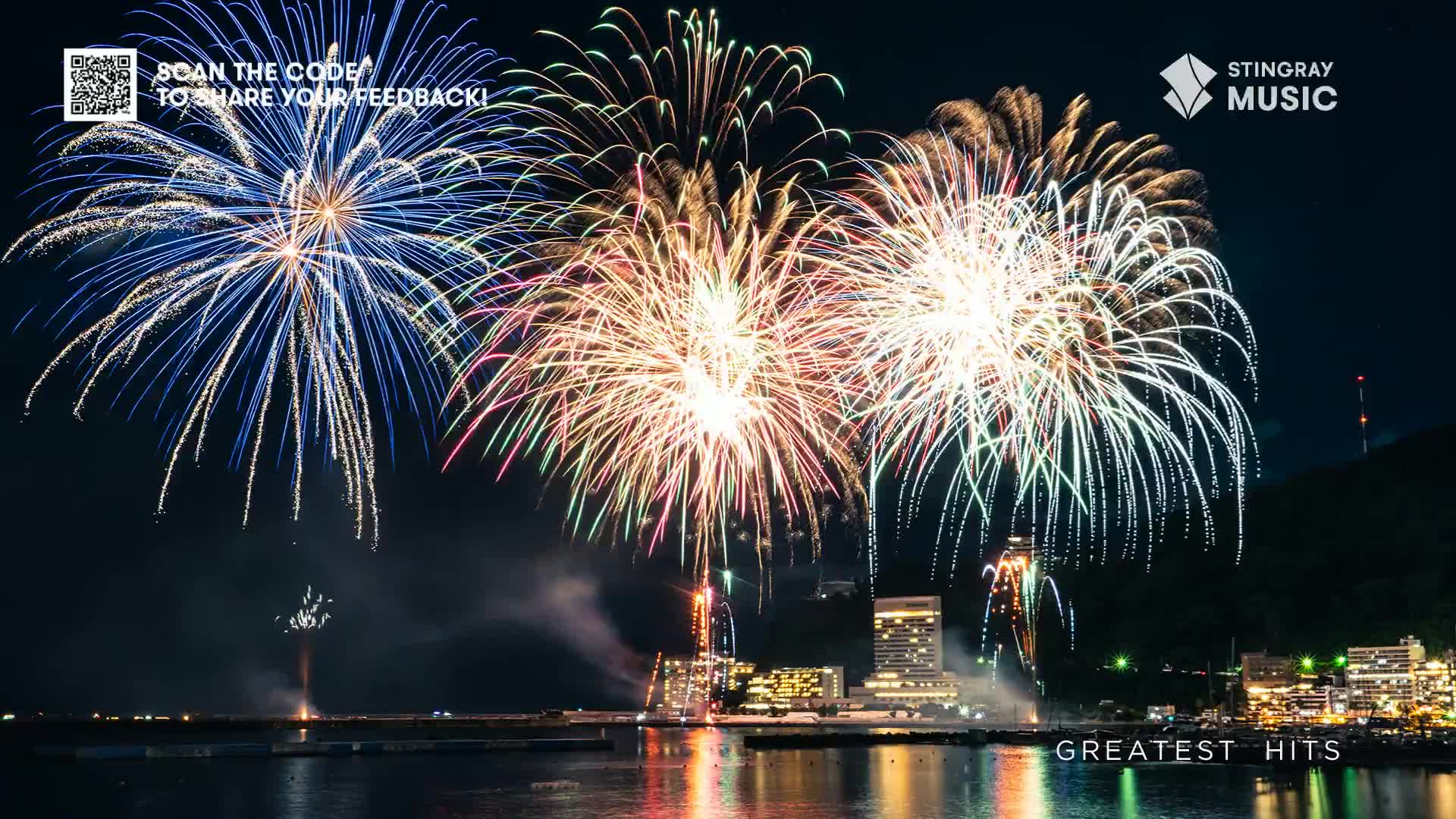 Bright bursts of fireworks erupt over the water, their trails of light reflecting in the dark surface. The scene is framed by a city skyline and a mountain, under a night sky that's perfect for a Stingray Holiday Hits celebration in Canada.
