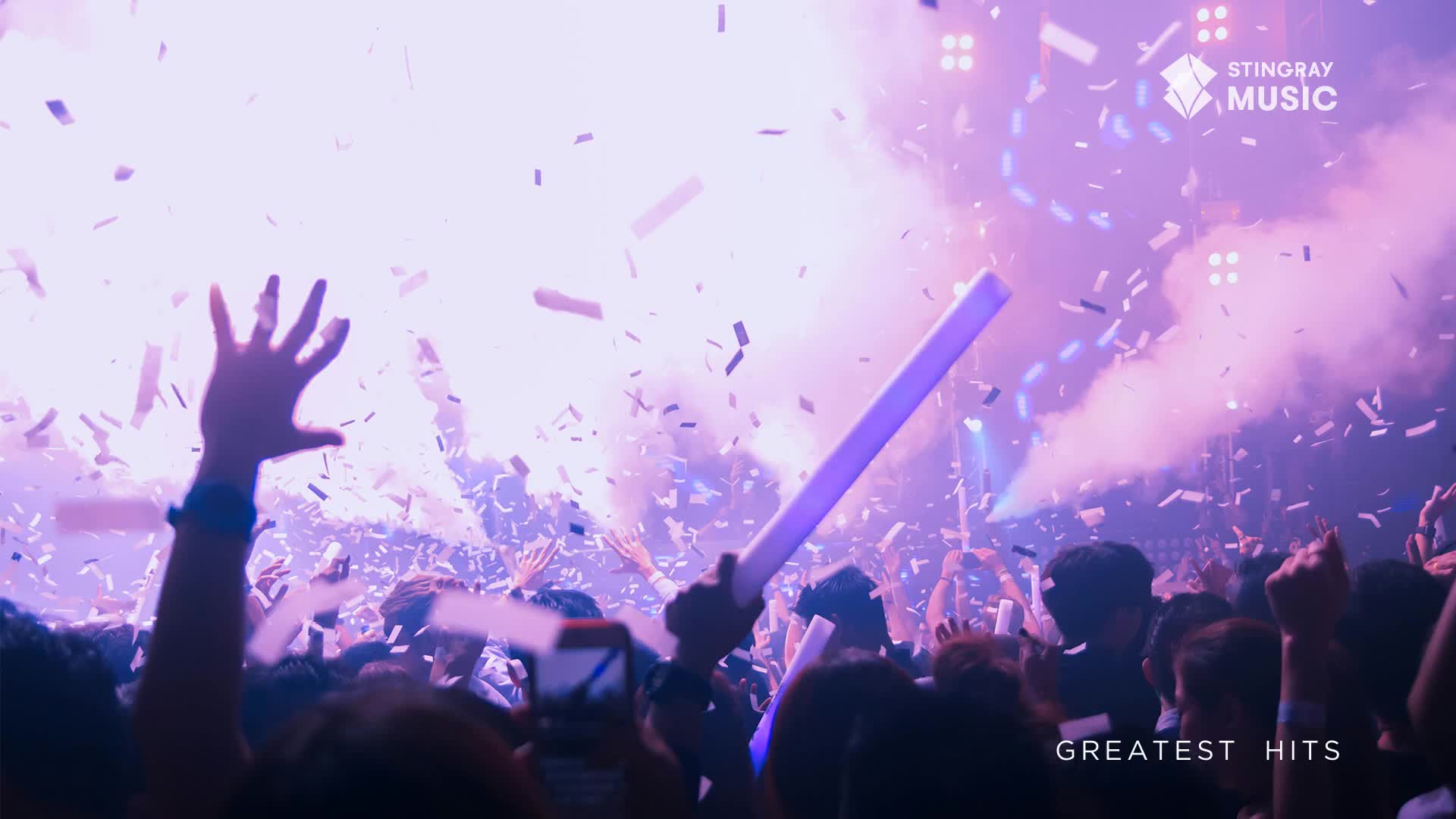 Confetti rains down on a crowd of people, their hands raised in the air. The stage lights are bright, and the air is thick with smoke.

