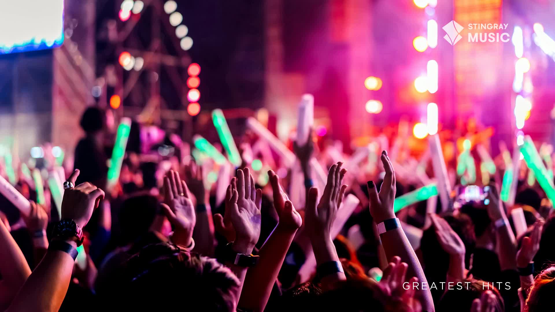 Arms and glowing green sticks are raised high, a sea of hands reaching toward the stage. The crowd is bathed in a wash of pink and purple light, likely from the Stingray Holiday Hits concert.
