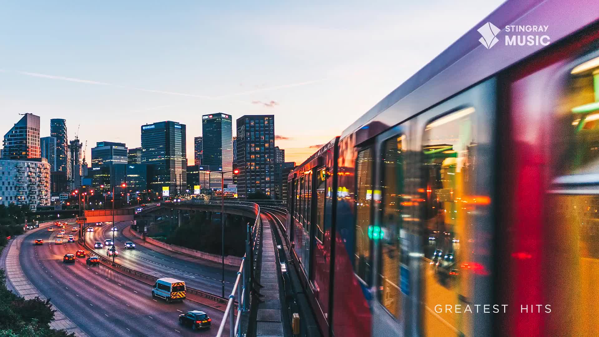 A red train rushes past, its windows reflecting the city lights and the evening sky.  Below, cars stream along a highway, and the Stingray Music logo sits in the upper right corner.
