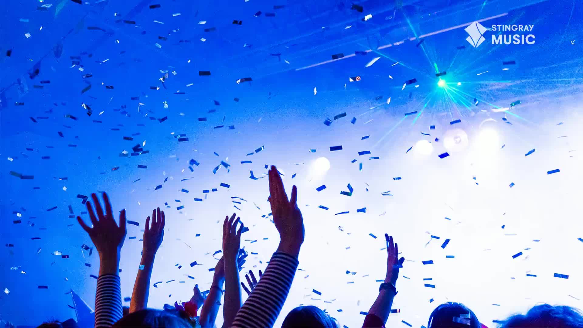Confetti flutters down, illuminated by bright stage lights, as arms reach up from the crowd. The blue and white lights create a festive atmosphere, perfect for a Stingray Holiday Hits concert in Canada.
