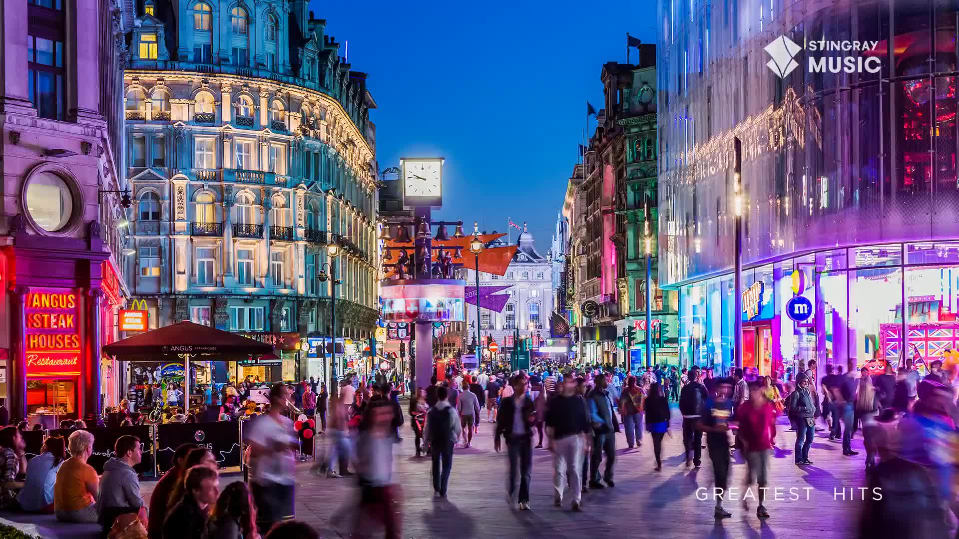 The street is packed with people, their forms blurred by movement under the glow of neon lights. Buildings rise up on either side, their facades illuminated in a mix of colors as the evening deepens.
